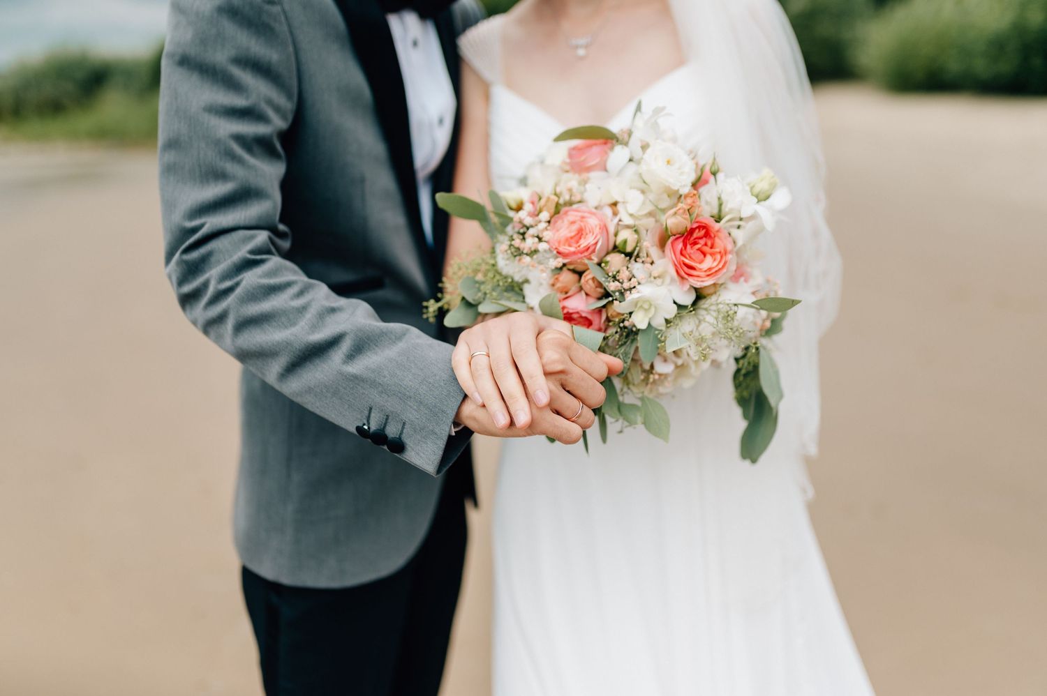 Close up of newlyweds holding coral and white wedding bouquet on beach.