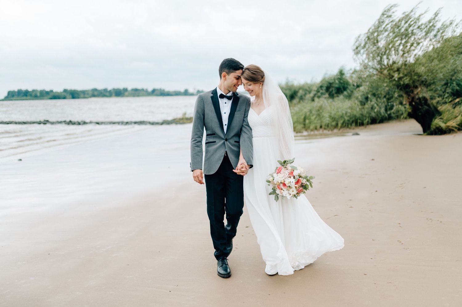 Couple walking along shoreline in formal wedding attire with bridal bouquet.