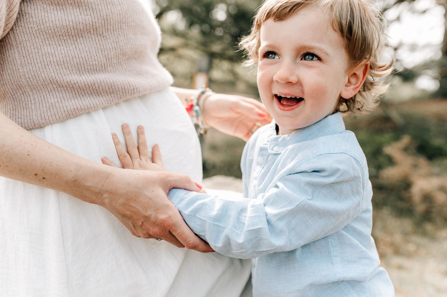A toddler in light blue clothing interacts playfully with a pregnant belly.