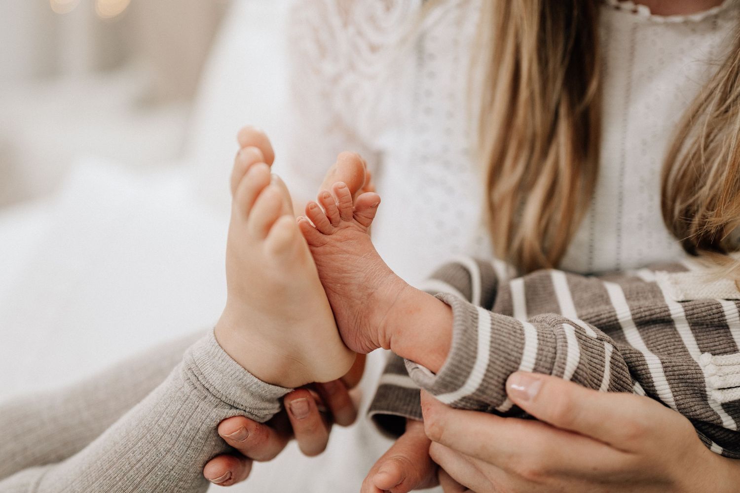 Baby's bare feet resting on caregiver's lap being held with caring hands in a cozy indoor setting.