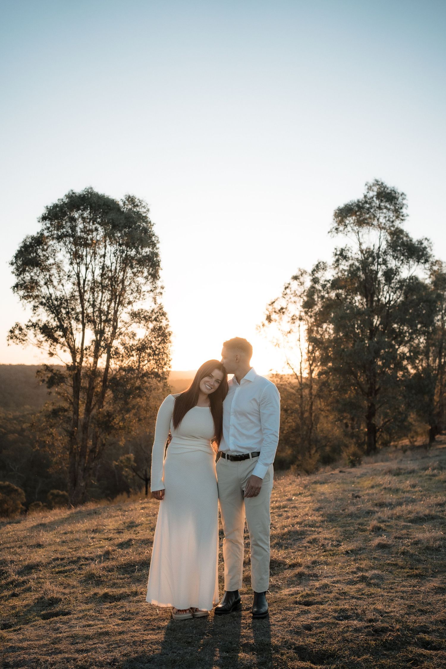 A couple embraces in formal attire during sunset on a hilltop with tall eucalyptus trees silhouetted against the sky.