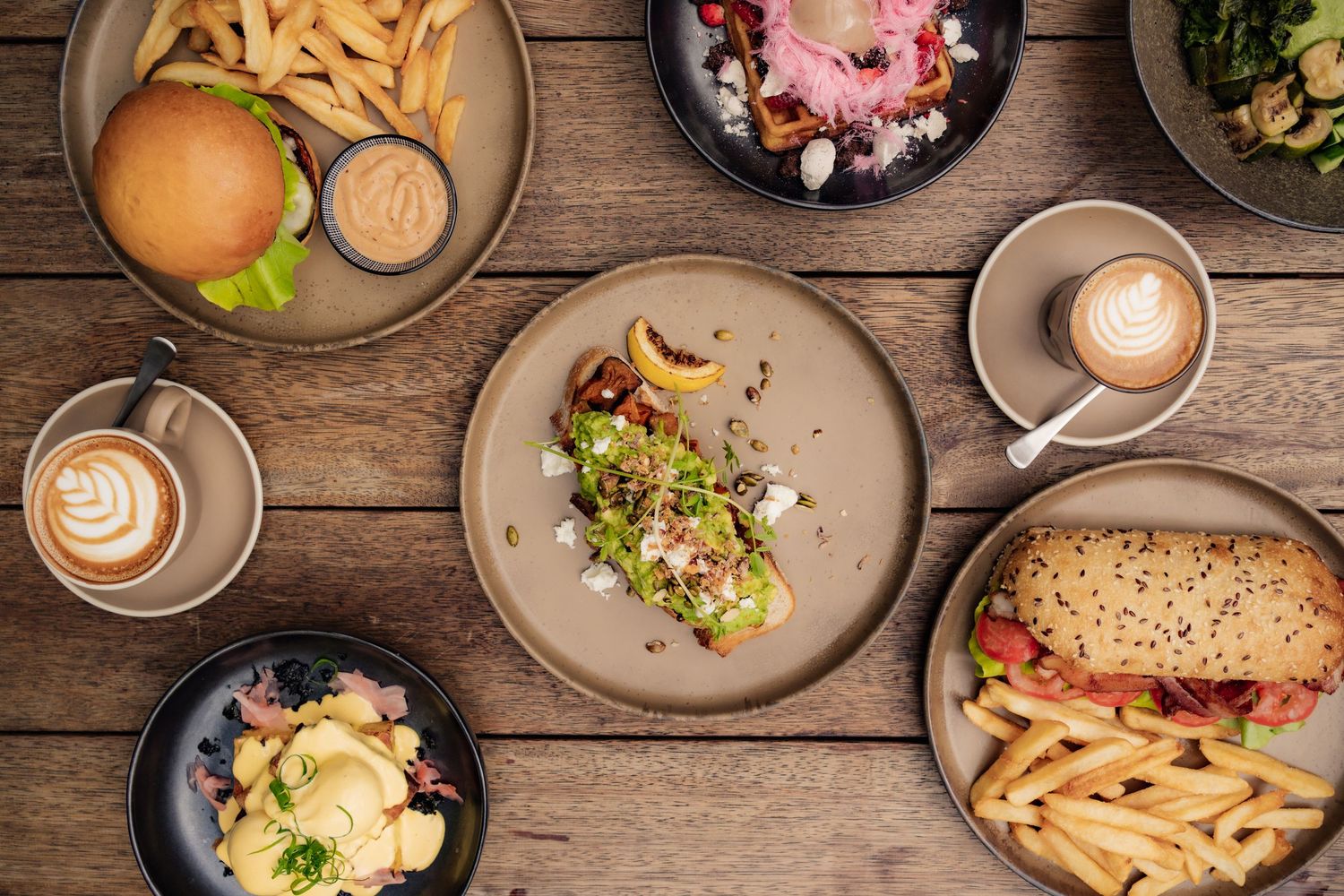 Overhead view of cafe brunch spread with burgers, french fries, coffee drinks and salads on rustic wooden table.