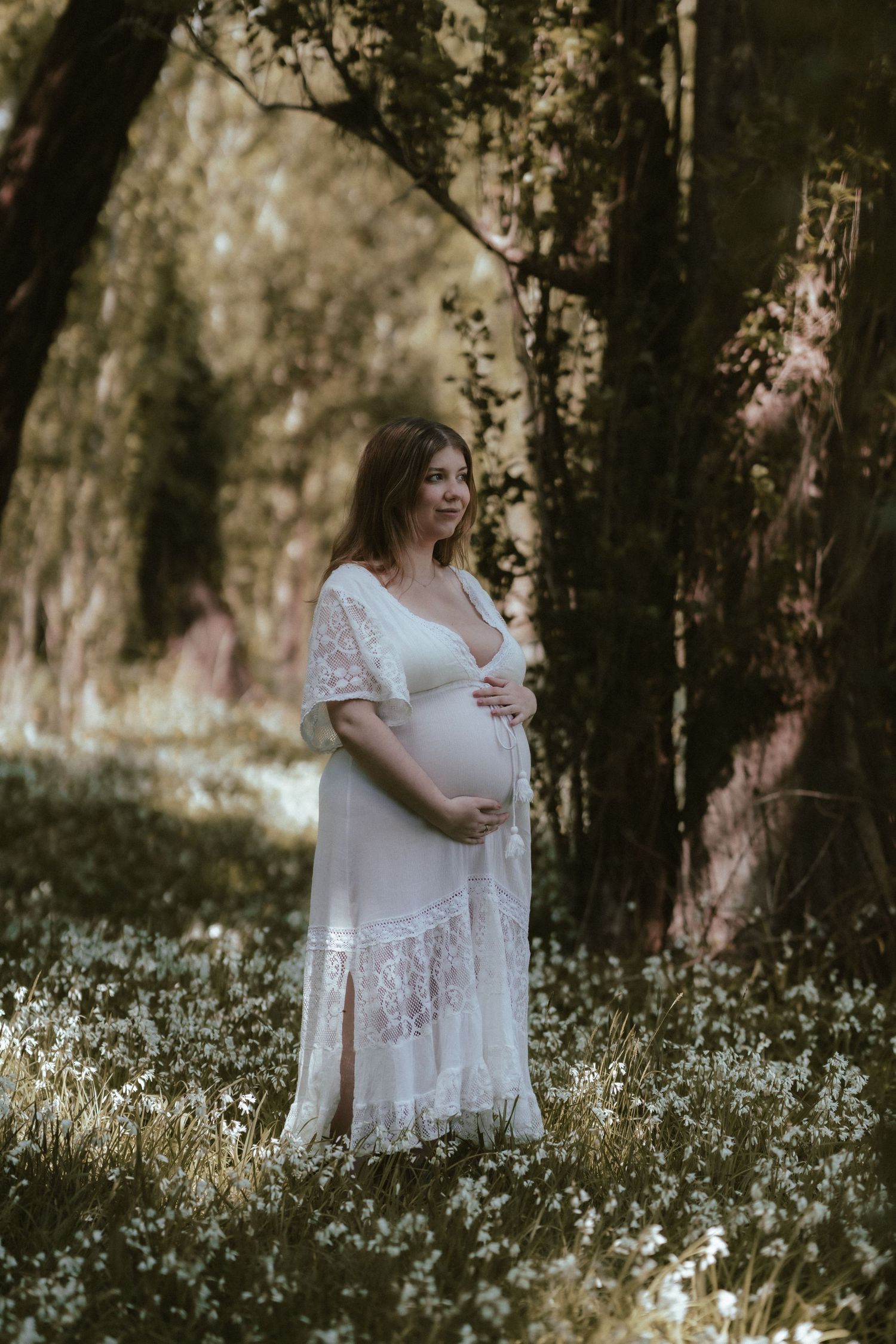 Ethereal maternity portrait in a white lace dress standing among wildflowers in dreamy sunlit forest setting.