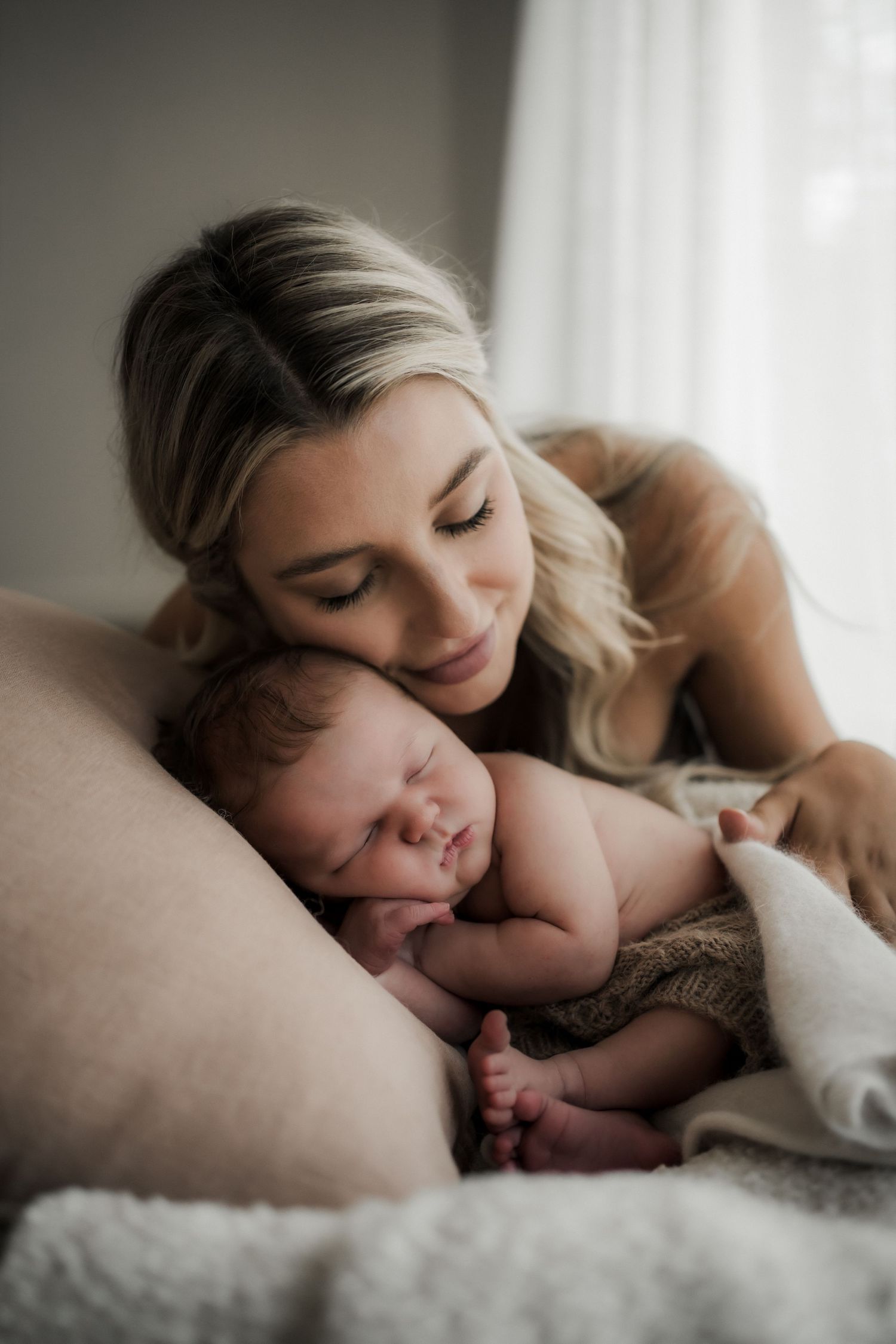 A tender moment captured as a mother and newborn baby snuggle together on soft bedding in natural window light.