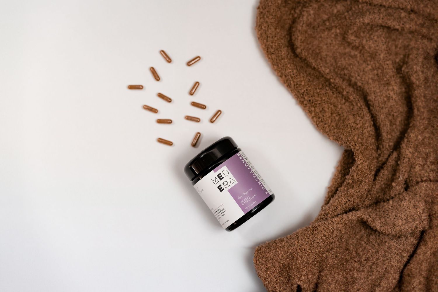 Purple vitamin supplement bottle with scattered capsules on brown textured blanket against white background.