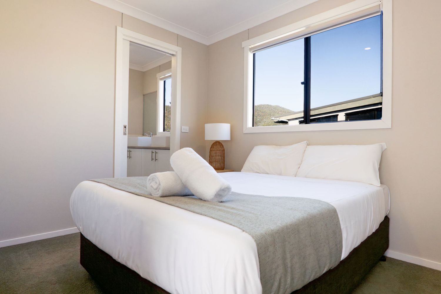 A modern minimalist bedroom with white bedding and linens features soft natural light streaming through windows.
