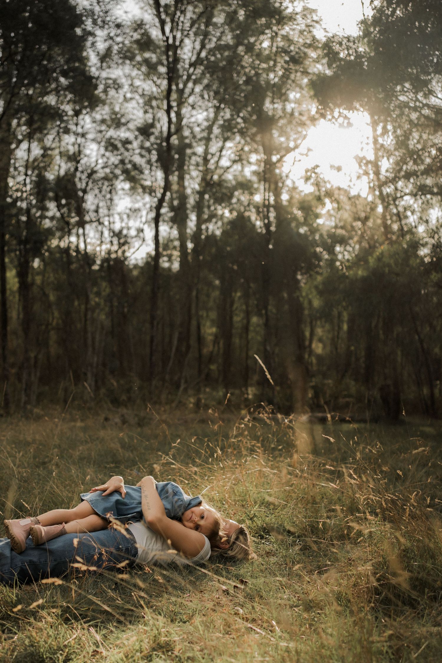 Two people lying together in tall grass during golden hour in a peaceful forest setting.