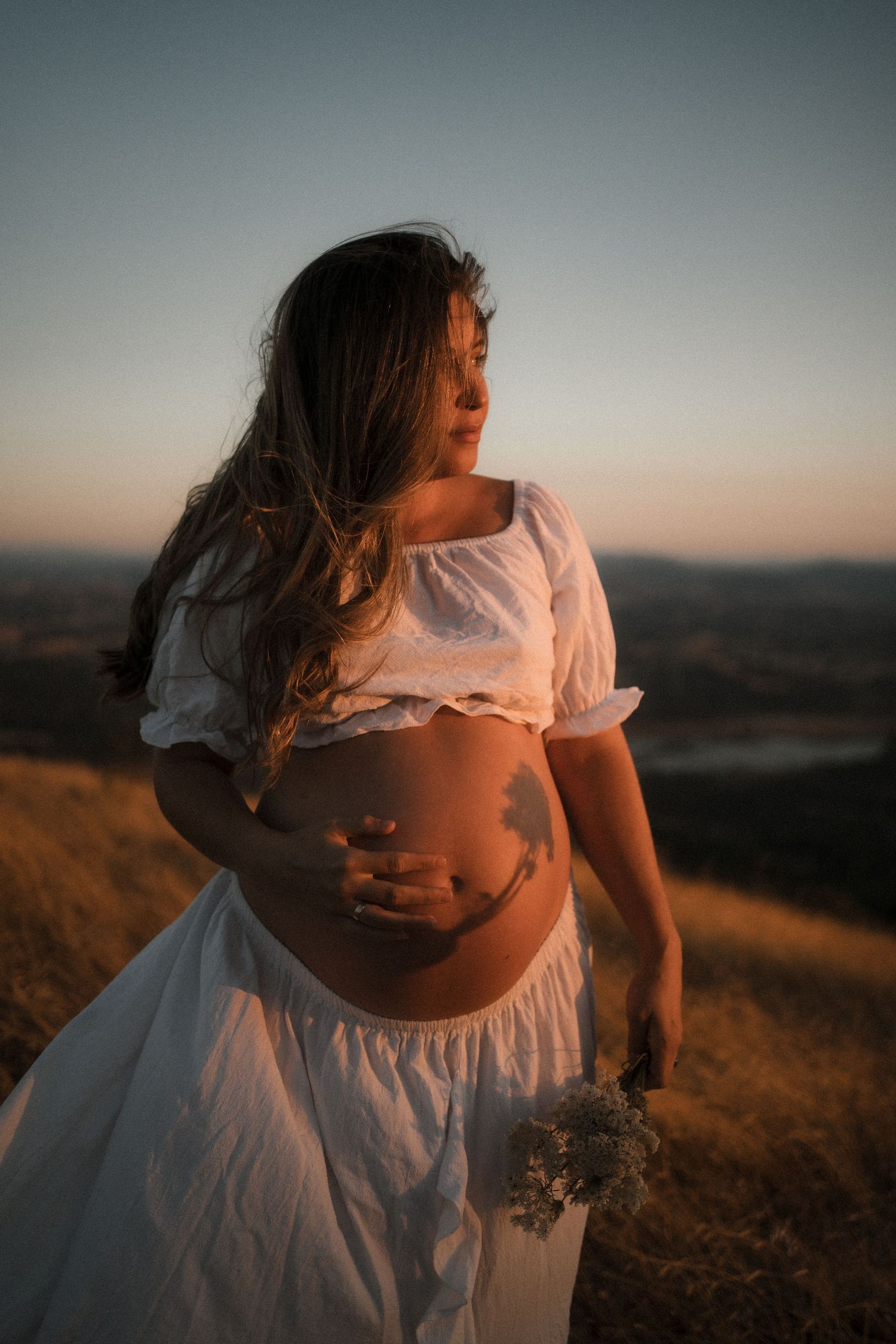 A pregnant woman in a white dress stands on a hill at sunset holding dried flowers in golden light.
