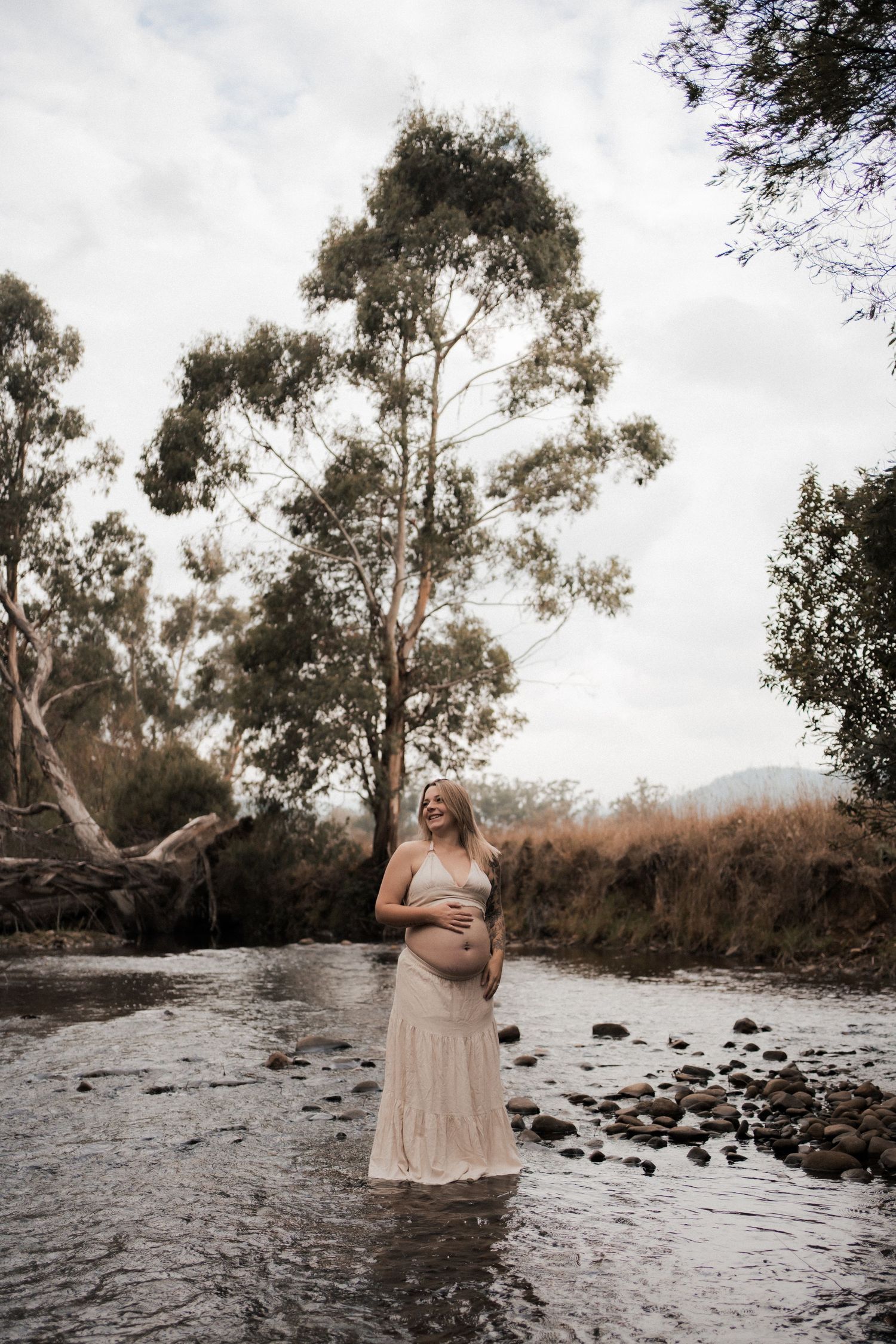 A figure stands in shallow river water wearing a long cream-colored dress amid eucalyptus trees and a moody sky.