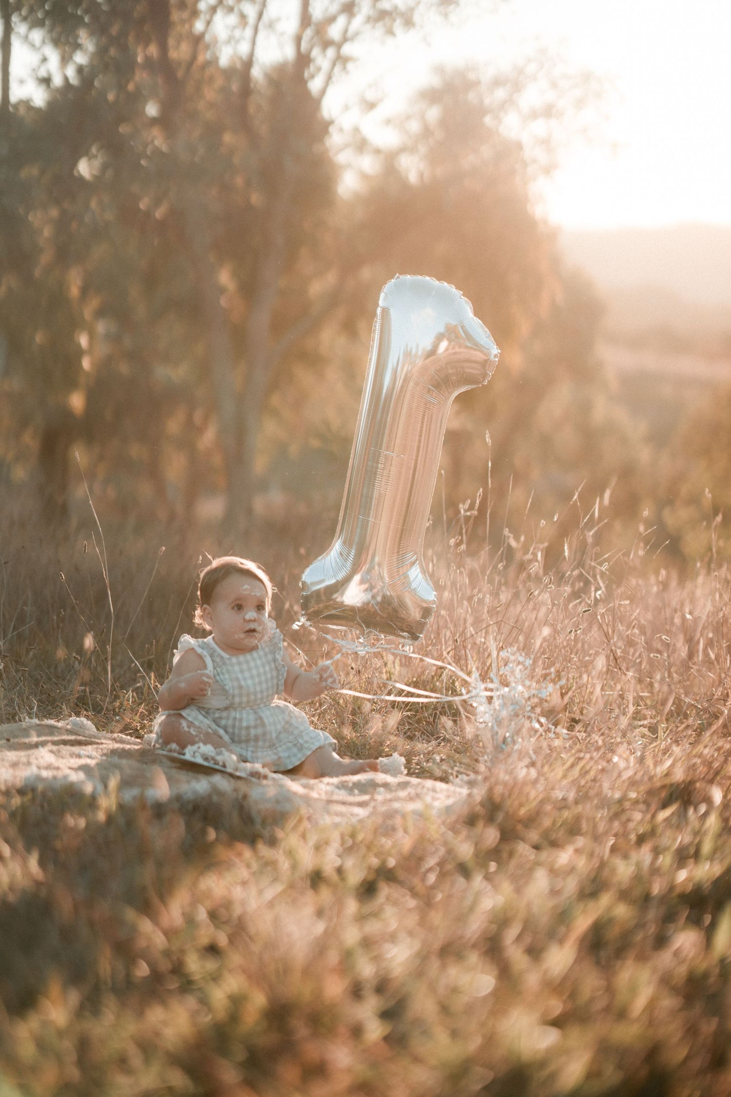 First birthday celebration outdoors at sunset with large silver number one balloon and baby sitting on blanket in field.