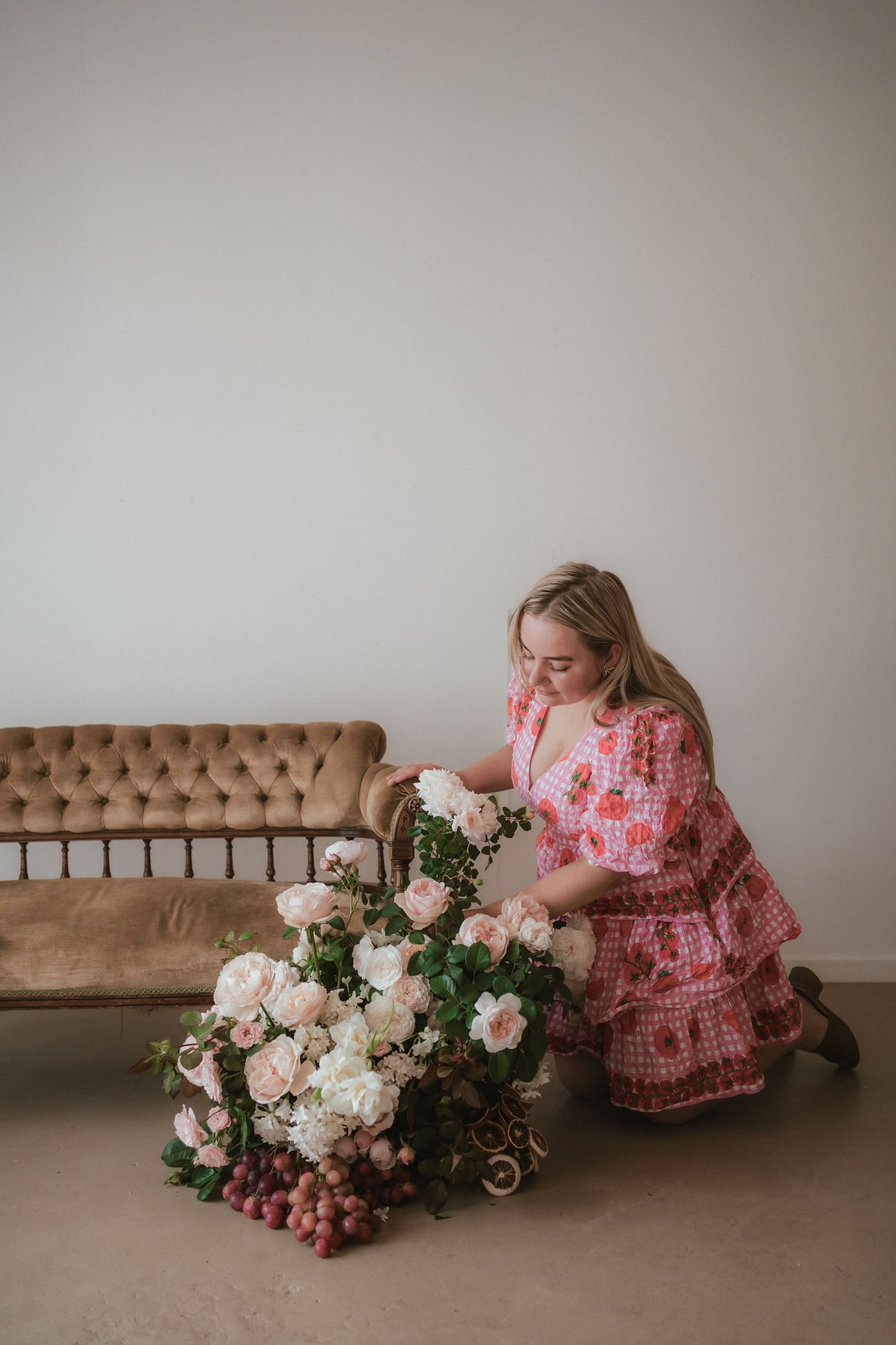 Woman in pink floral dress arranges elegant white and blush peony flower arrangement next to brown vintage sofa.