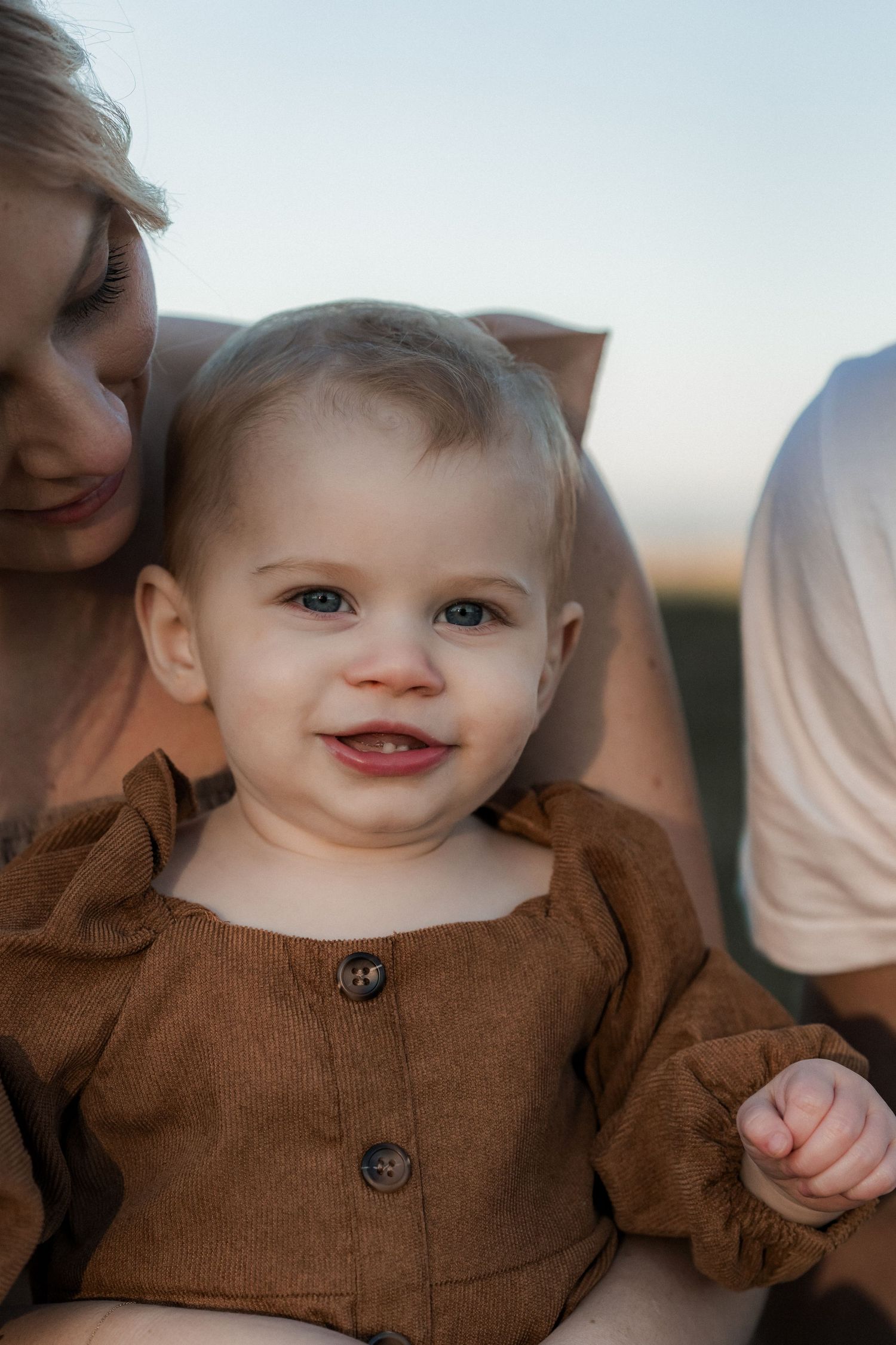 Toddler wearing brown button-up shirt smiles while sitting outdoors with family on a sunny day.