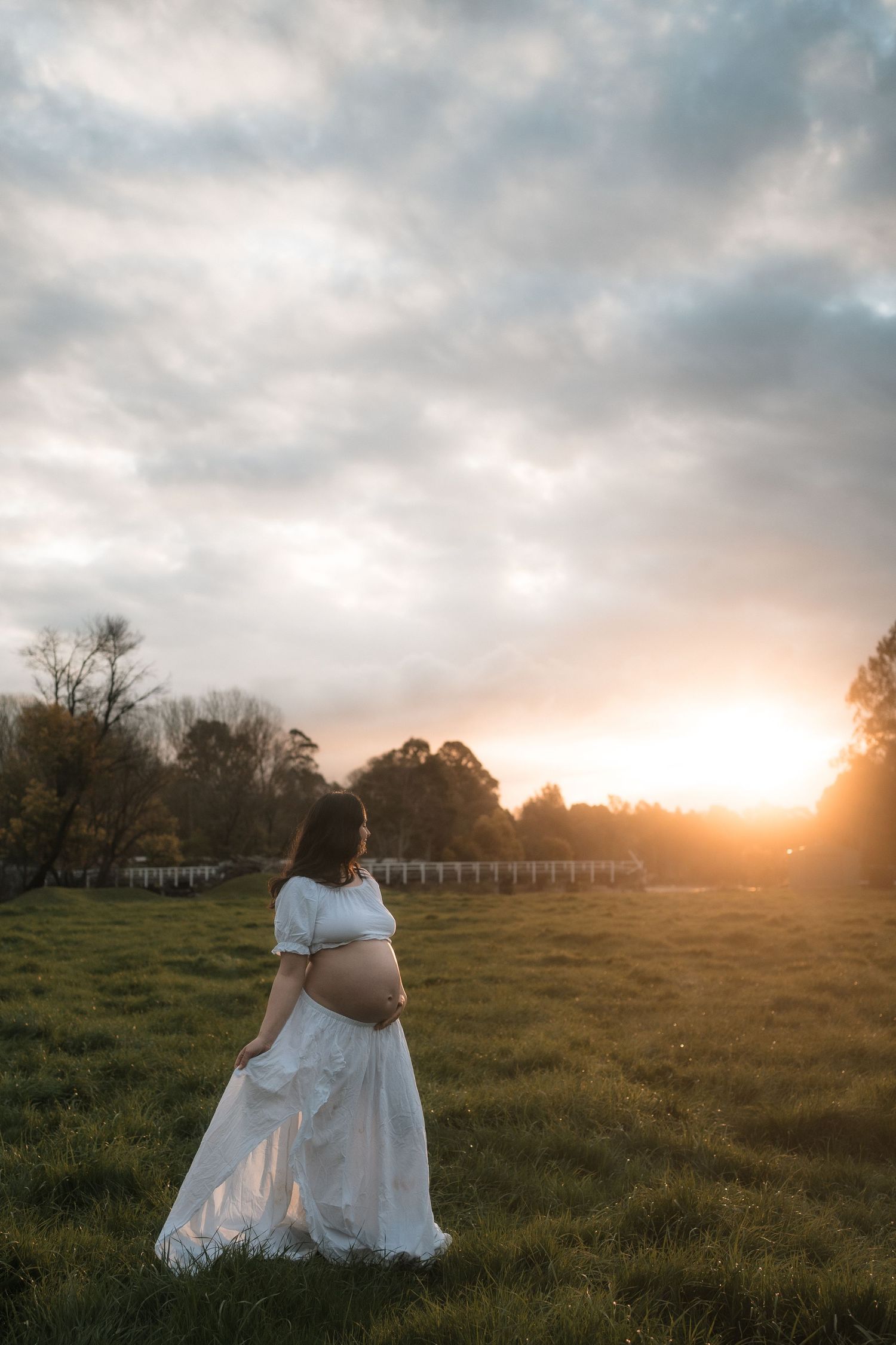 Pregnant woman in white dress standing in green field at golden sunset, cradling baby bump outdoors.