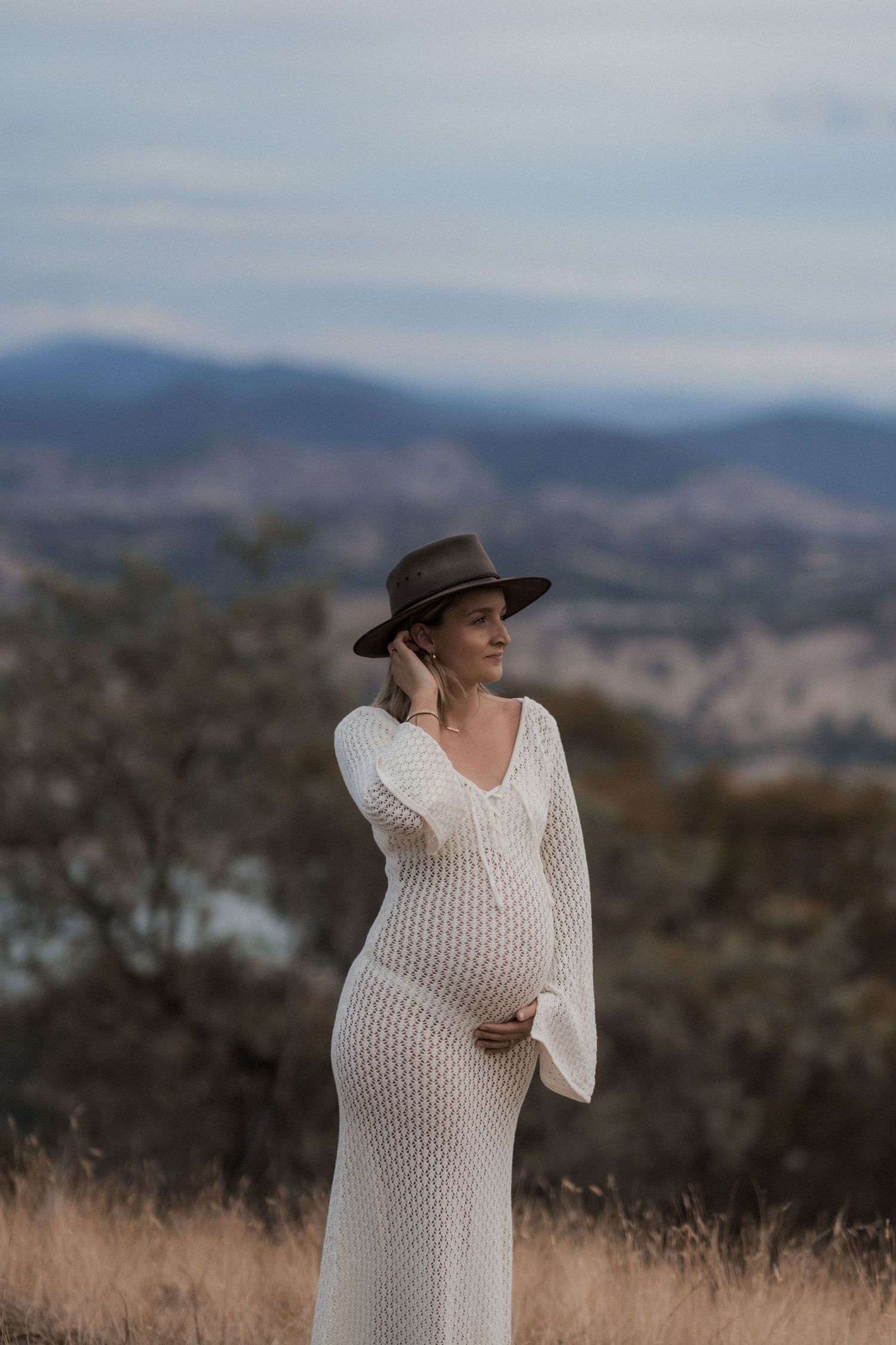 A maternity portrait featuring a woman in white knit dress and hat against mountain backdrop.