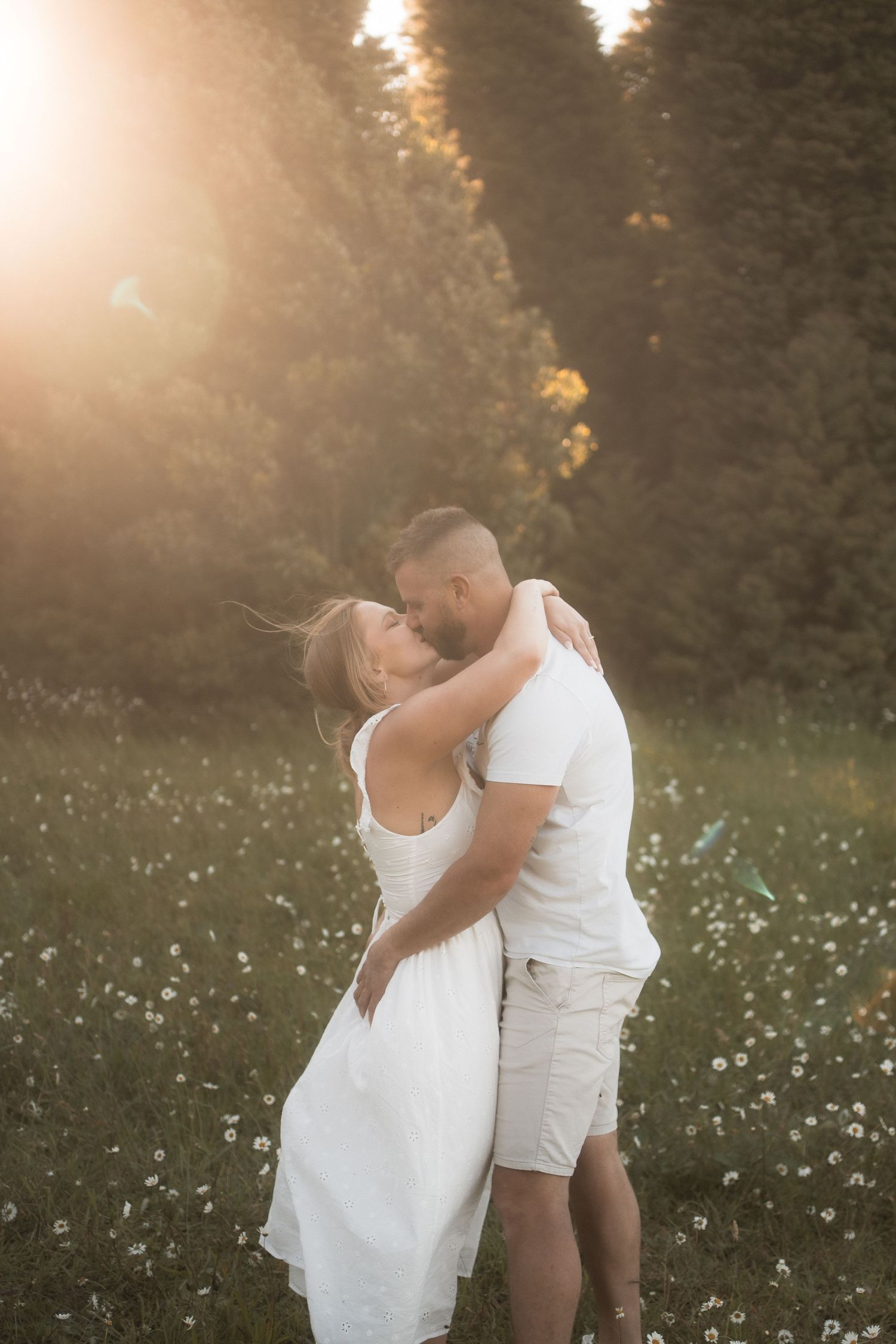 A couple in white outfits embraces romantically in a sunlit meadow filled with wildflowers at golden hour.