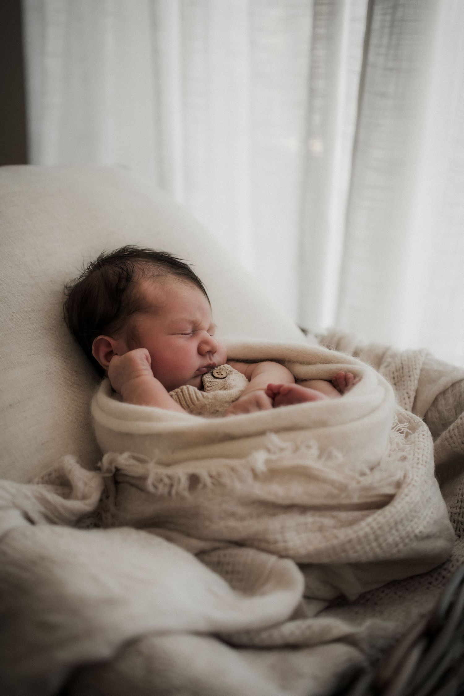 A sleeping newborn baby wrapped in cozy cream-colored blankets rests peacefully against white cushions.