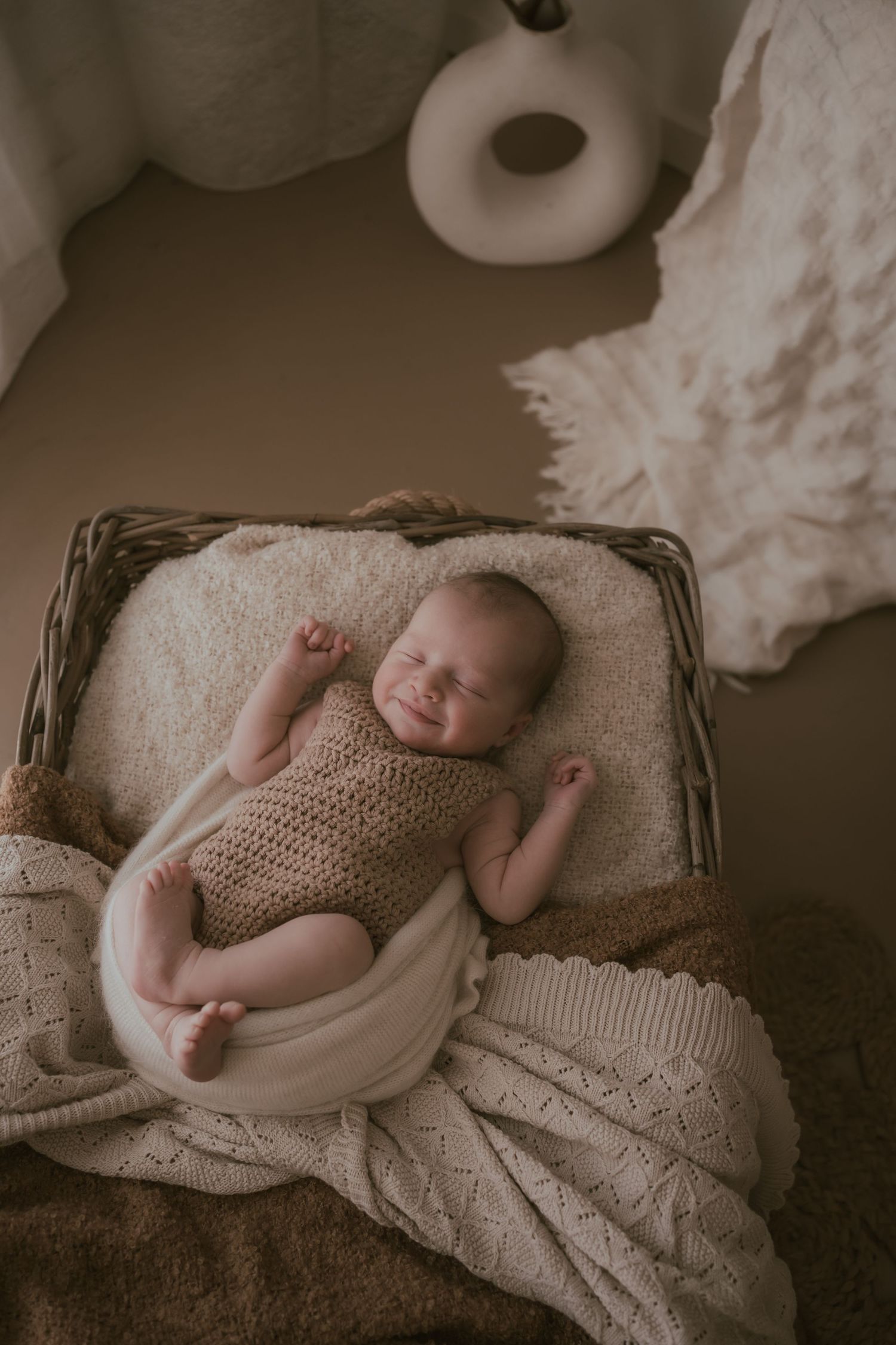 Sleeping newborn wrapped in cozy knitted beige romper lies peacefully in woven basket with soft textured blankets.