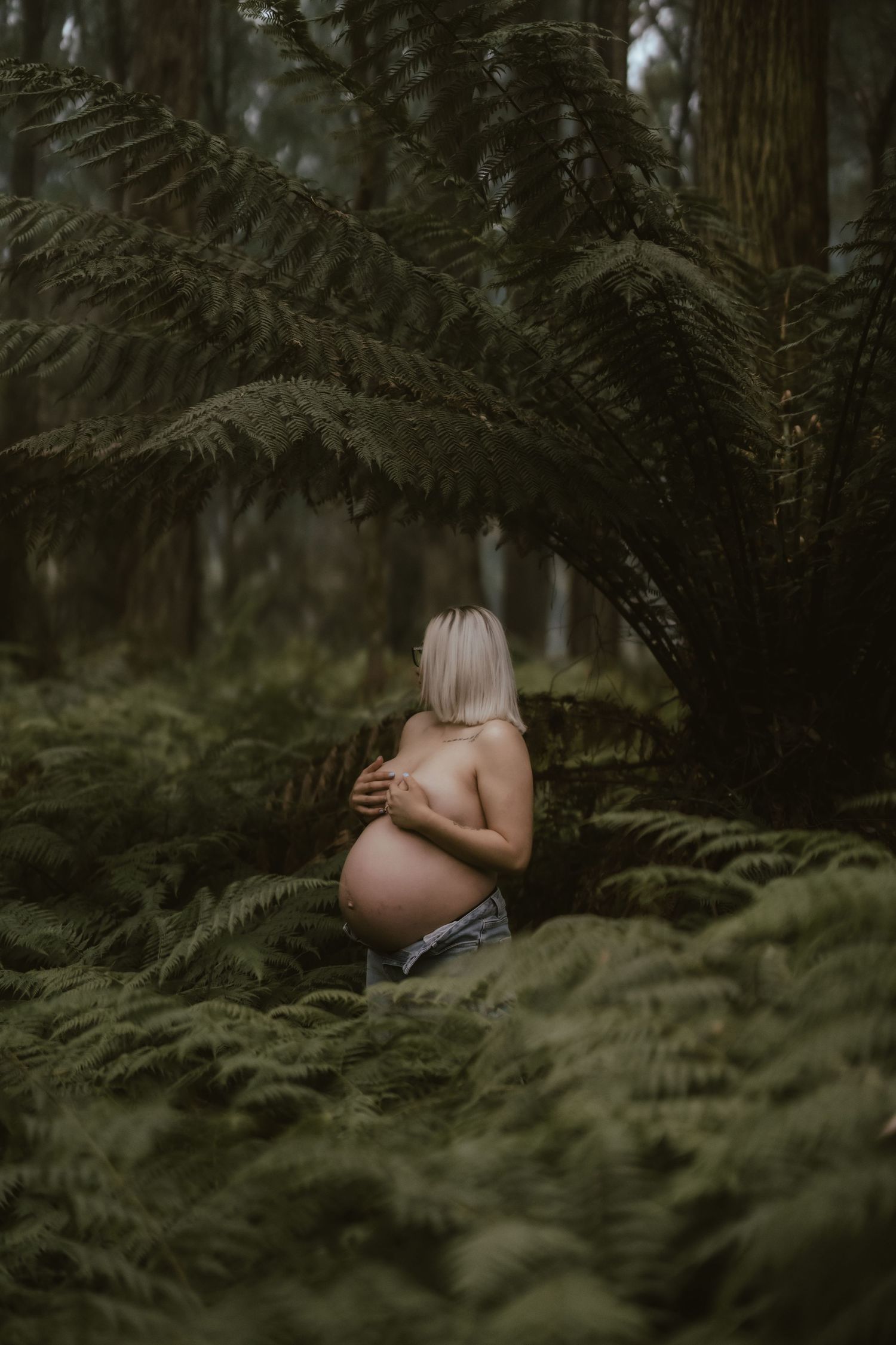Dark moody maternity photo shows pregnant figure crouching among lush green ferns in Pacific Northwest forest.