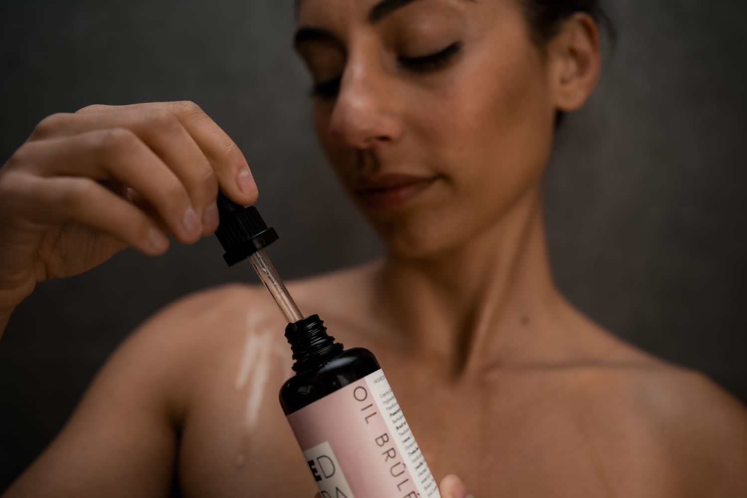 Person holds dropper of pink skincare product serum against dark background while demonstrating application technique.