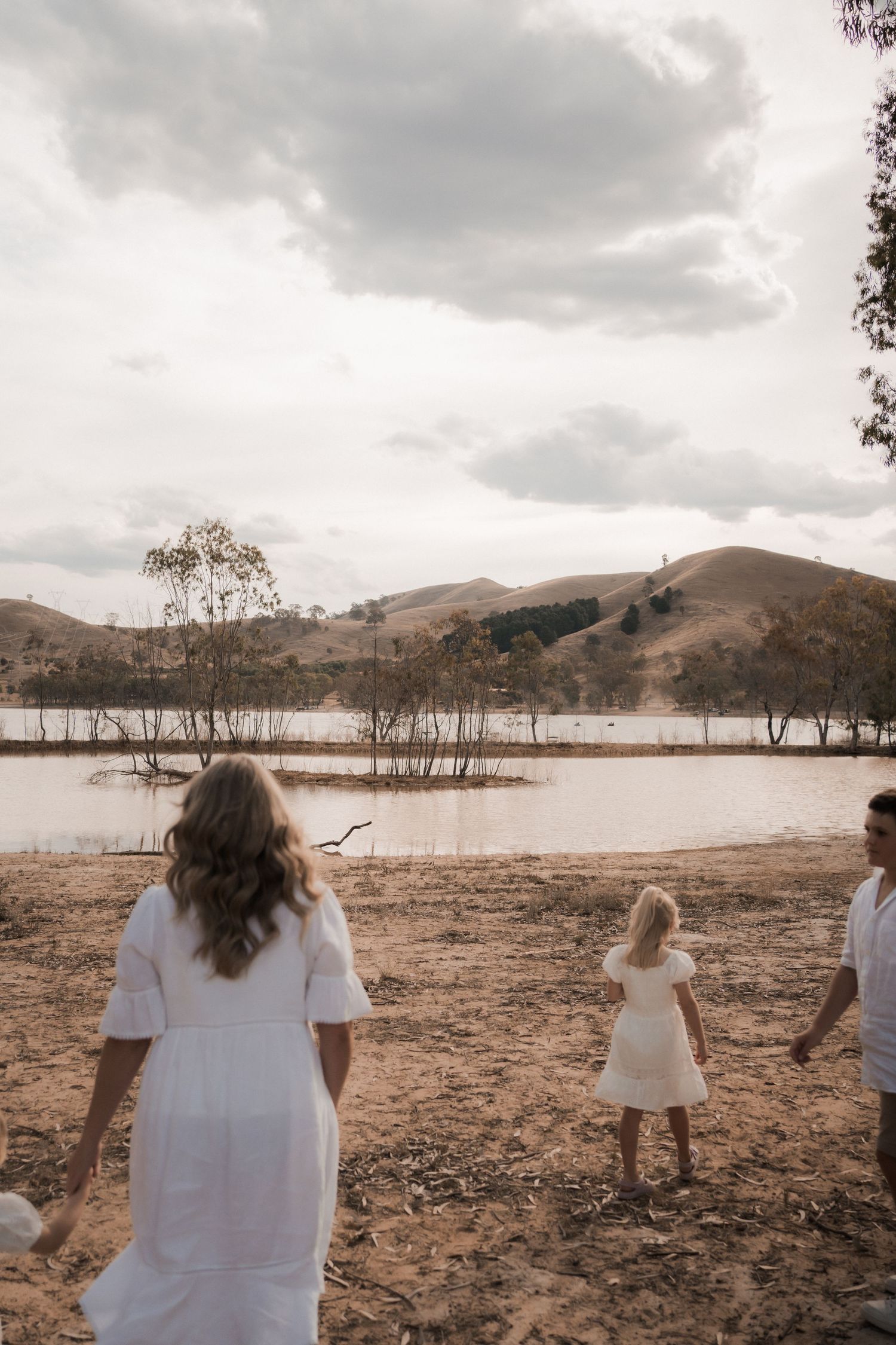 People in white clothing stand near a lake surrounded by hills and bare trees under cloudy skies.