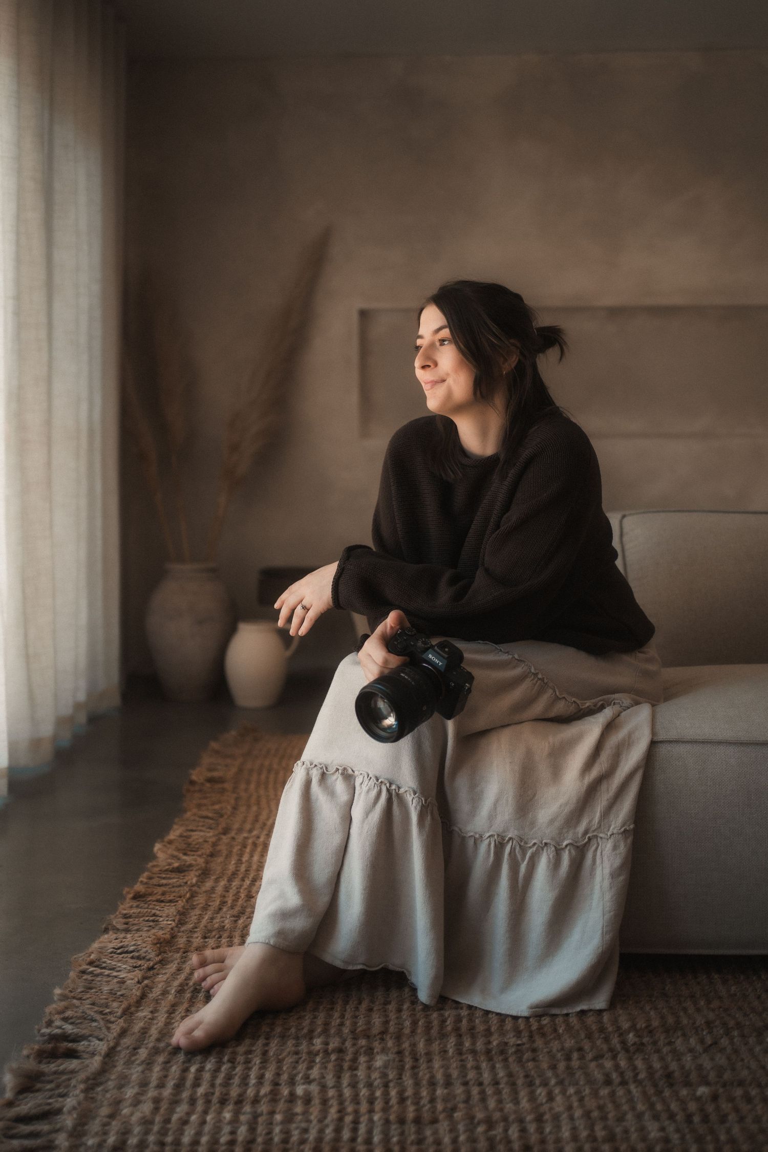 Woman in a dark sweater and linen skirt sits barefoot on a sofa holding a camera in a neutral room with dried pampas, vases and a jute rug.