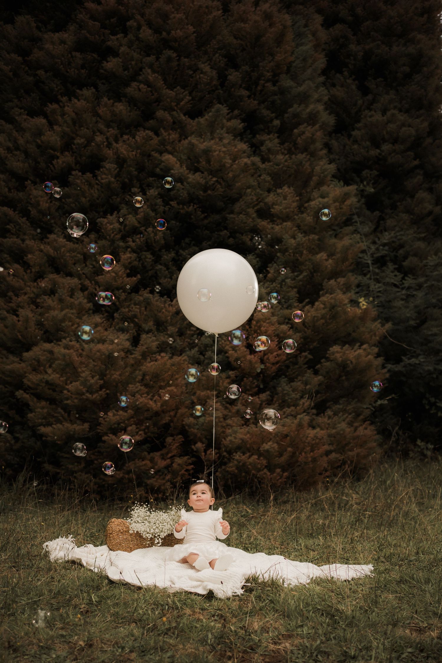 A white balloon and soap bubbles float around a blanket on grass against a dark forest background.