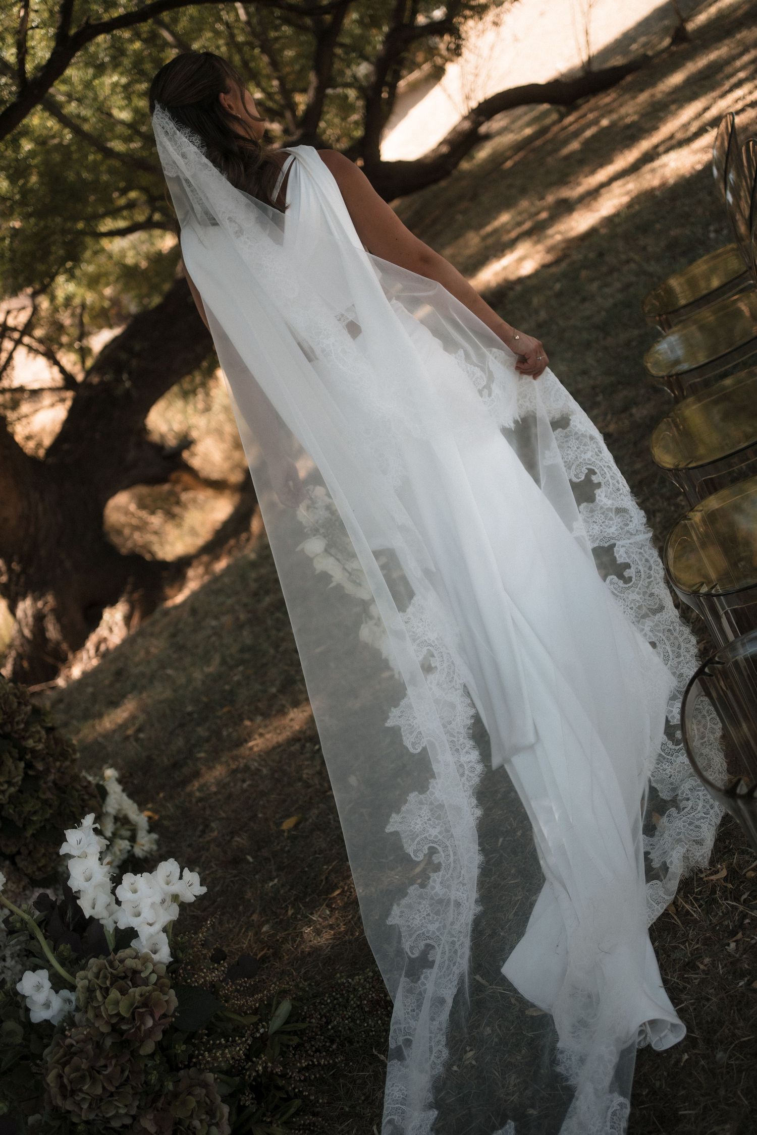 A flowing white wedding dress with intricate lace details is photographed in a natural outdoor forest setting.