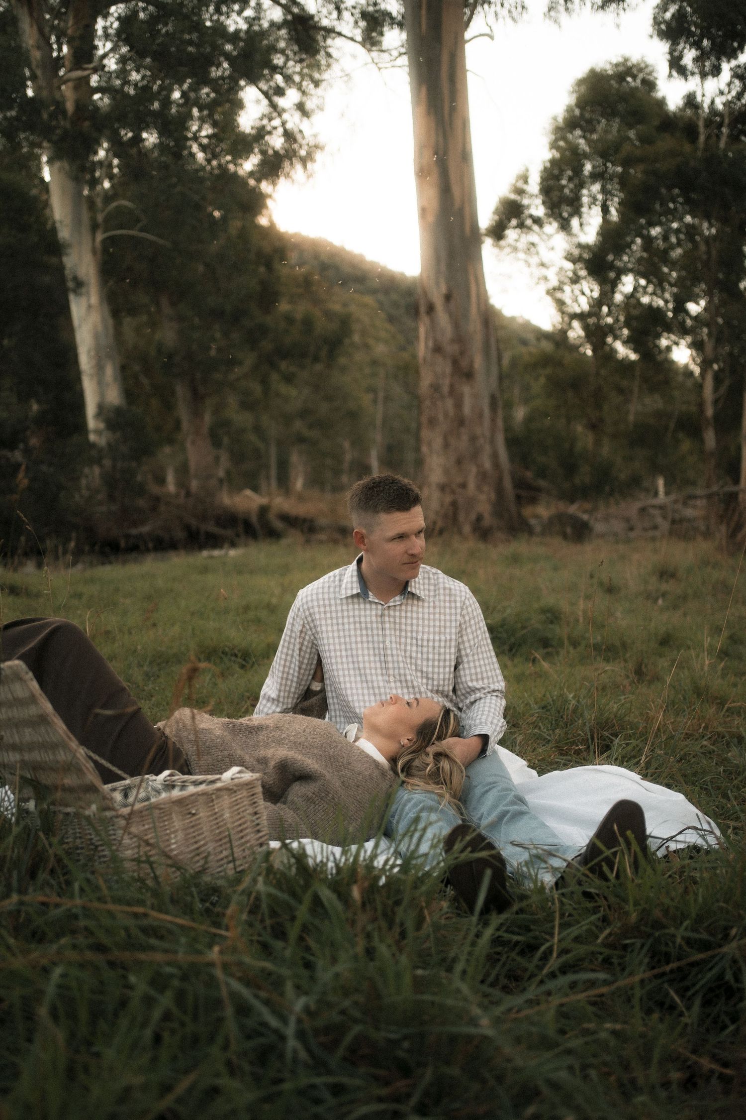 Couple enjoying a romantic outdoor picnic on grass, woman resting head on mans lap, surrounded by tall eucalyptus trees.