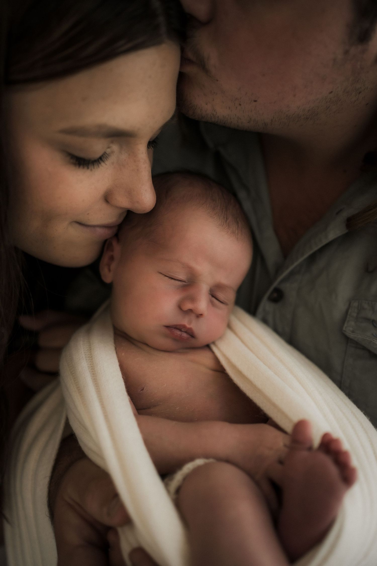 Intimate family portrait showing parents snuggling with their sleeping newborn baby wrapped in soft white fabric.