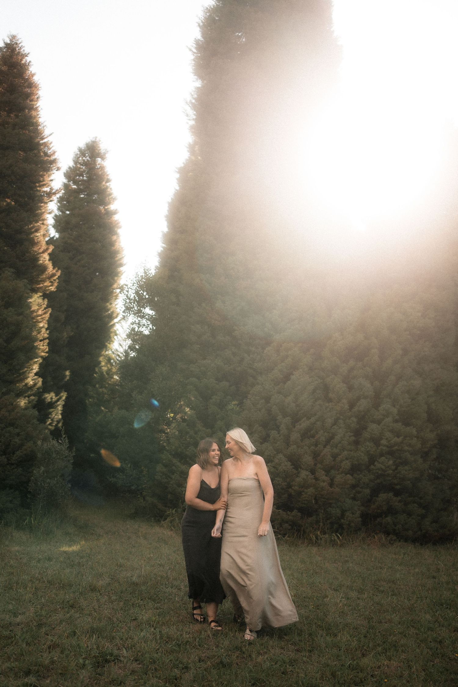 Two people in elegant dresses share a tender moment in a misty forest clearing at sunset.