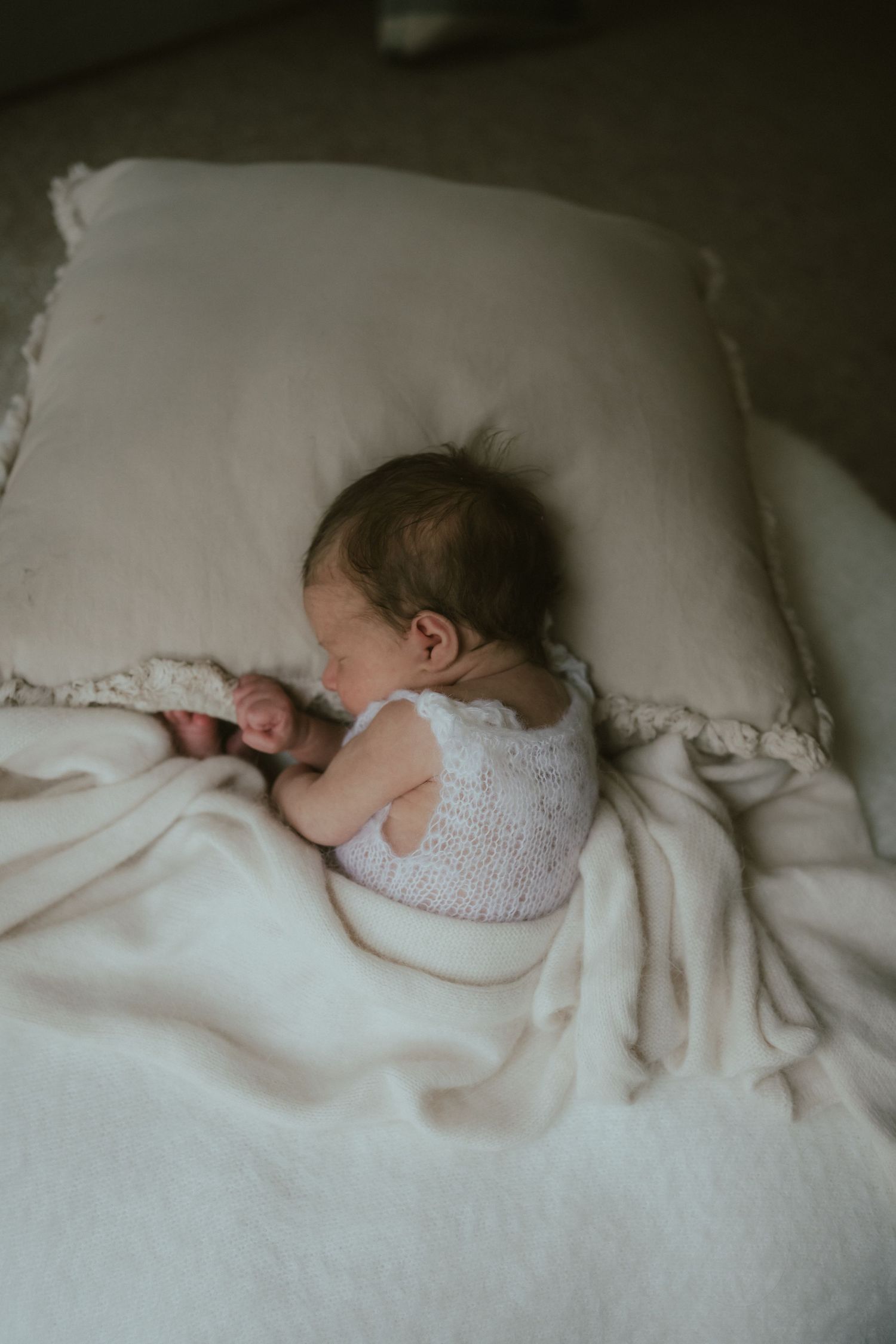 A sleeping baby wearing a white knit top rests peacefully against a white pillow and blanket in soft lighting.