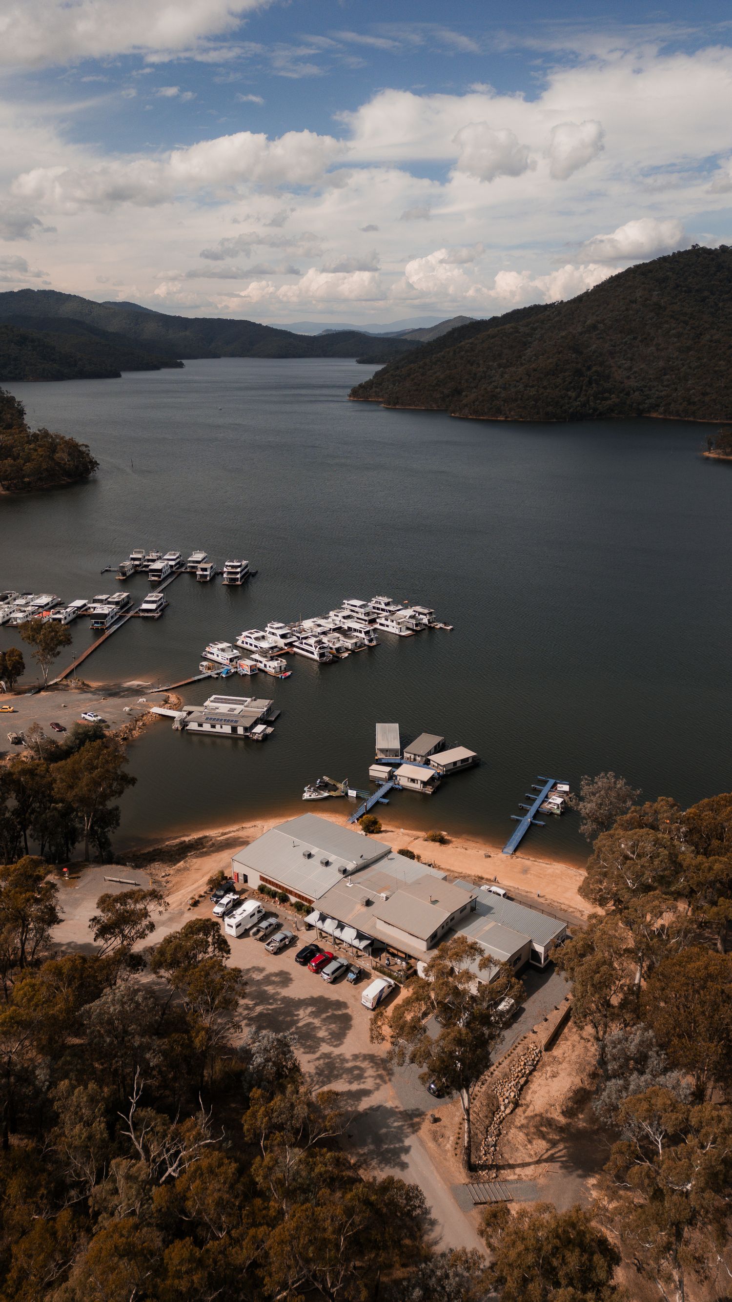 Aerial views of a marina with white houseboats docked at Lake Eildon in Australia, surrounded by forested hillsides.