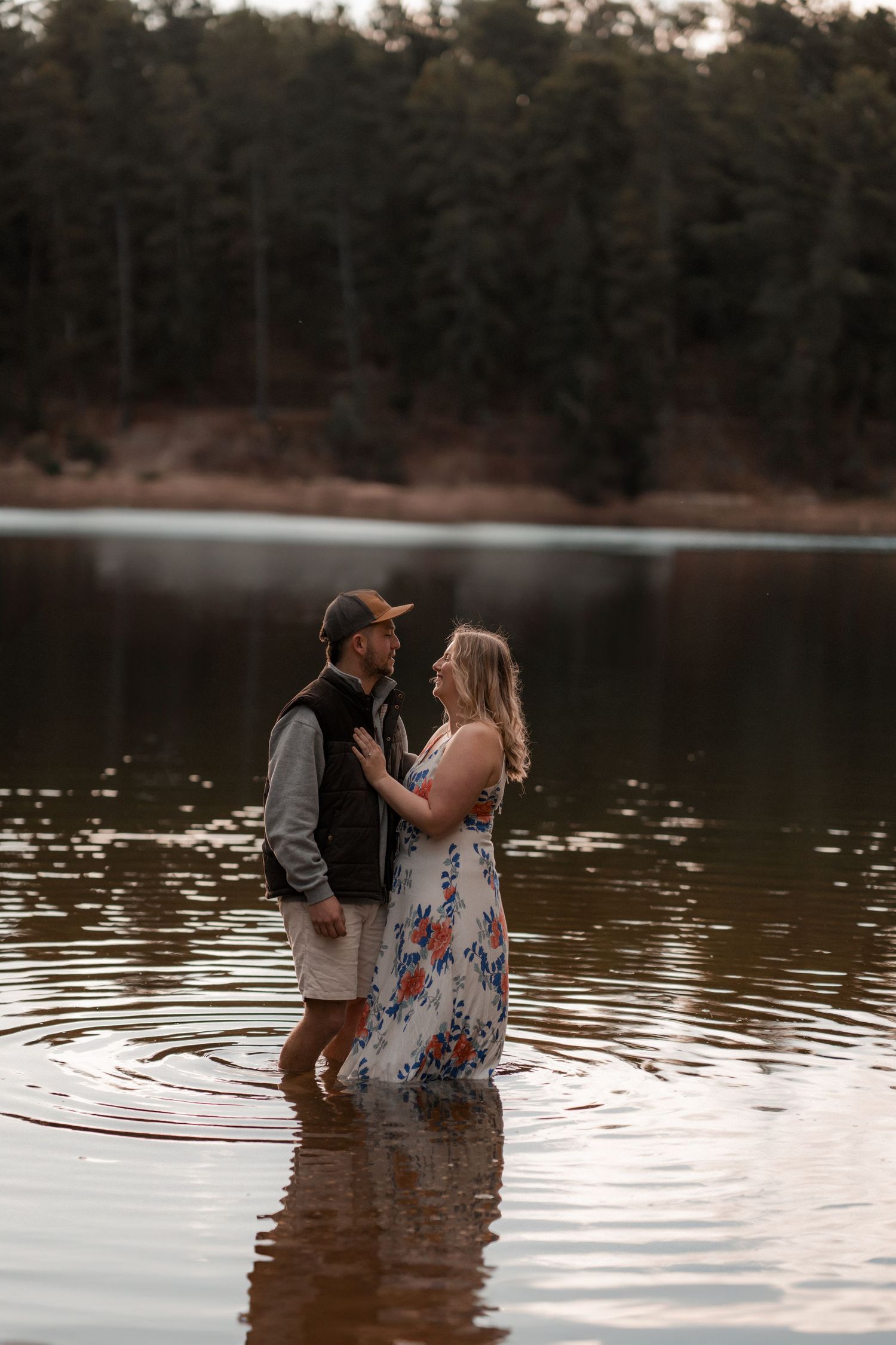 A romantic couple embraces while standing in shallow lake water at sunset with pine trees in background.