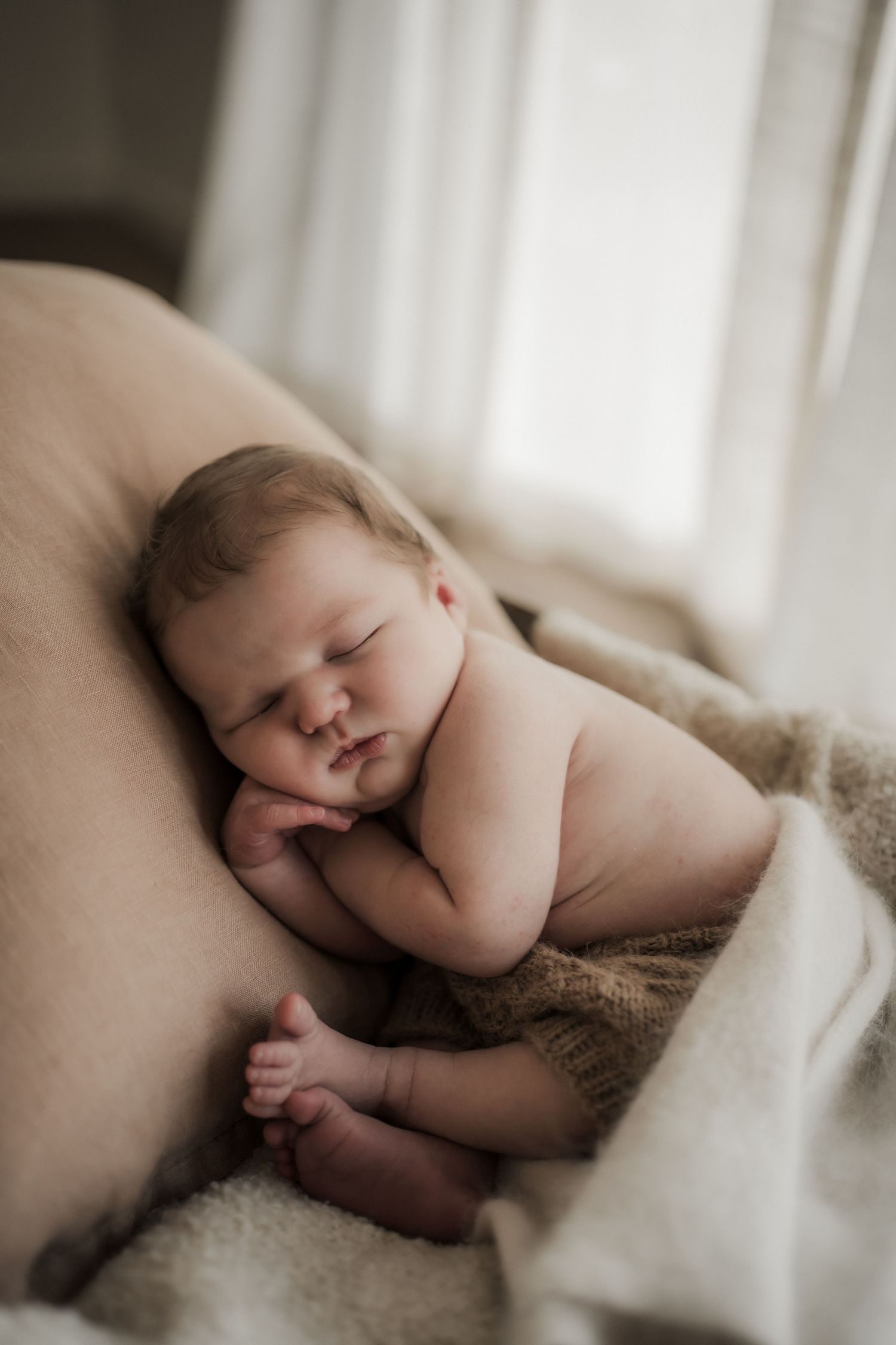 A peaceful sleeping newborn baby lies on soft brown fabric in natural window light.
