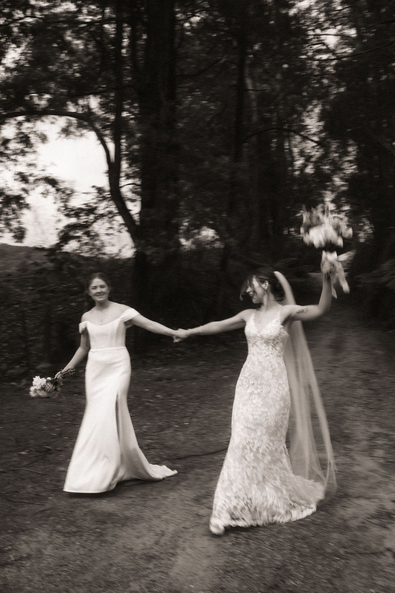 Two people in elegant white wedding dresses dance and twirl together in a natural outdoor setting with trees in the background.