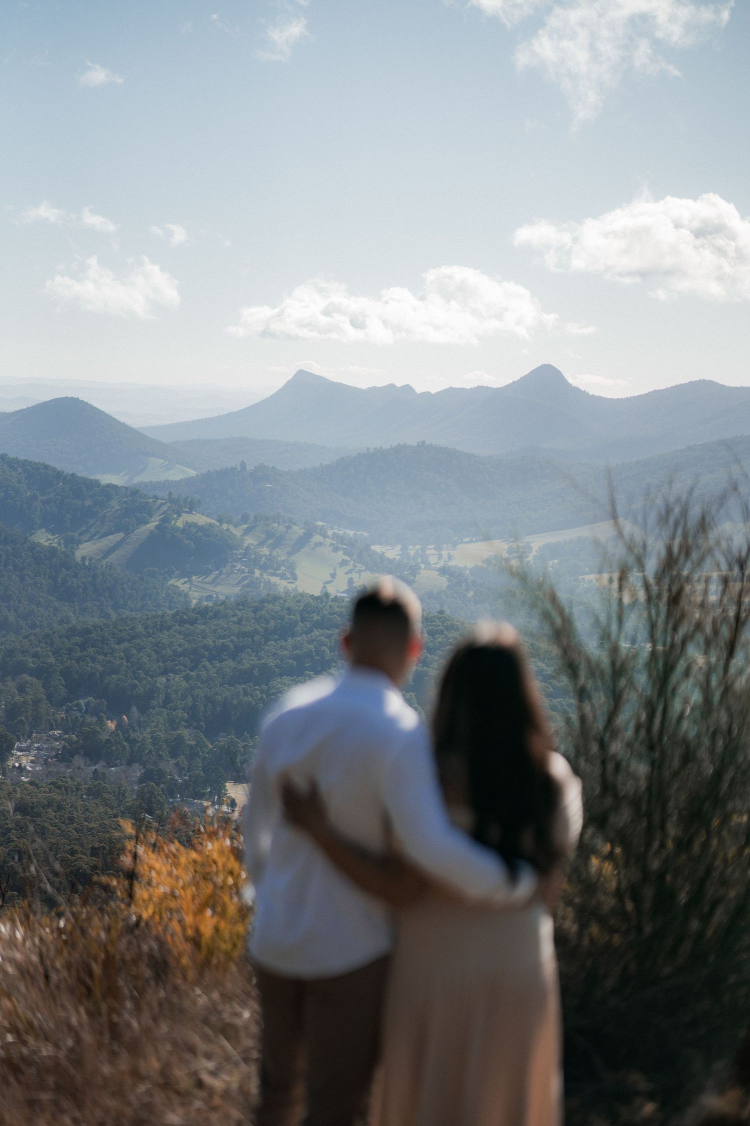 A couple embraces while overlooking a scenic mountain landscape with peaks and valleys under a partly cloudy sky.