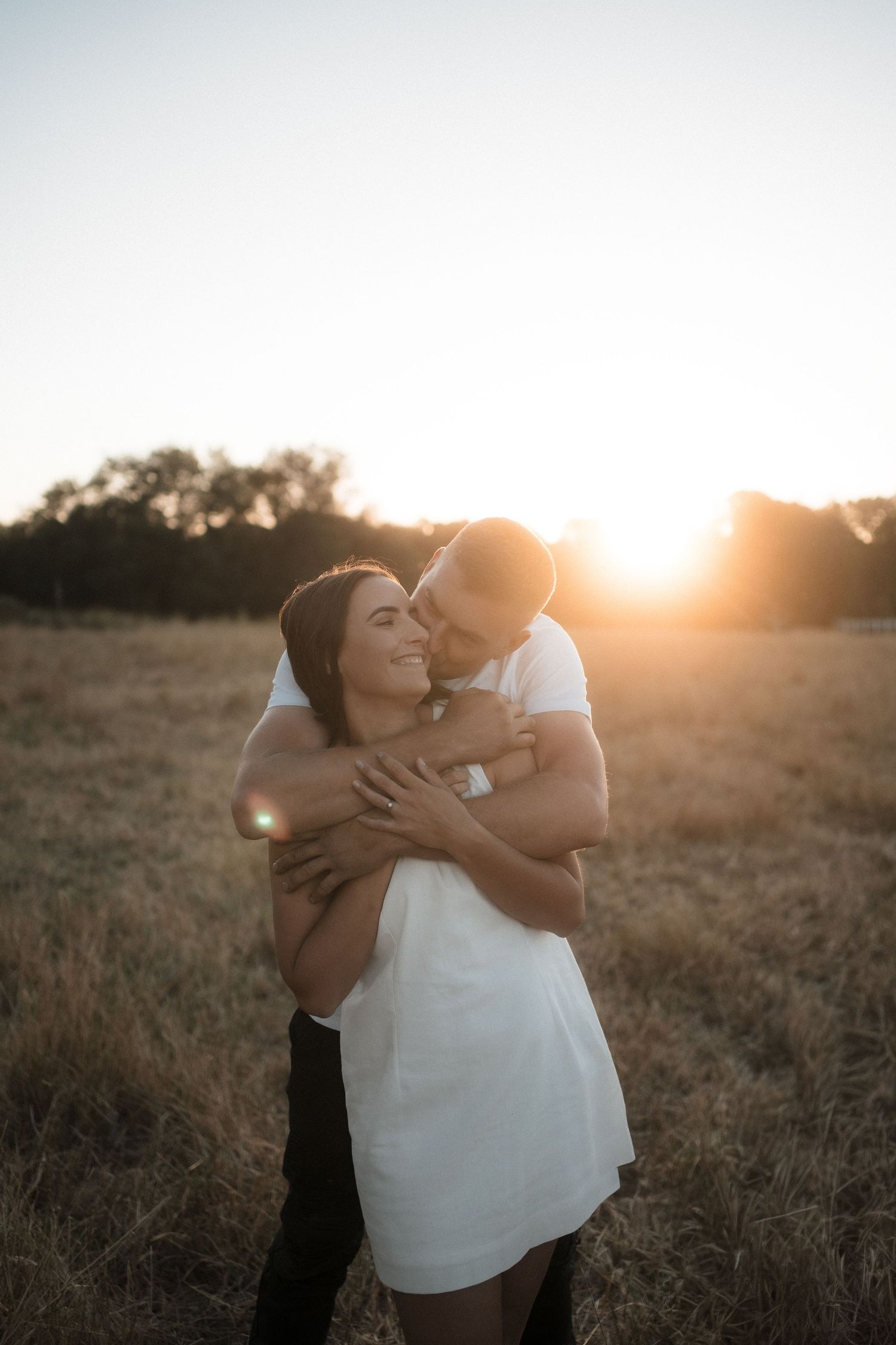 Couple embracing in golden field at sunset, woman smiling as man kisses her cheek in romantic outdoor portrait.