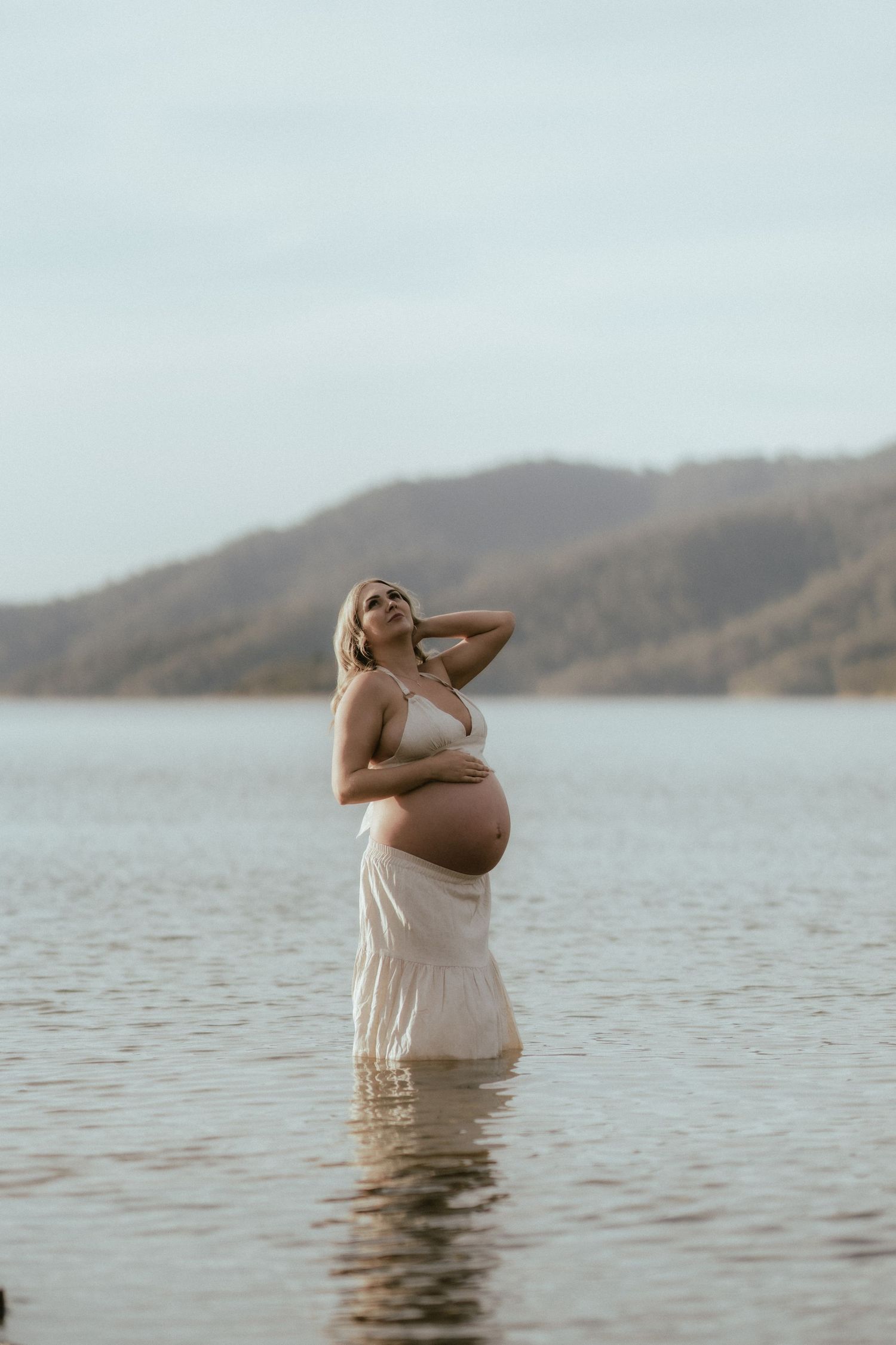 An expectant person stands in calm lake waters wearing a flowy white dress against a mountainous backdrop at dusk.