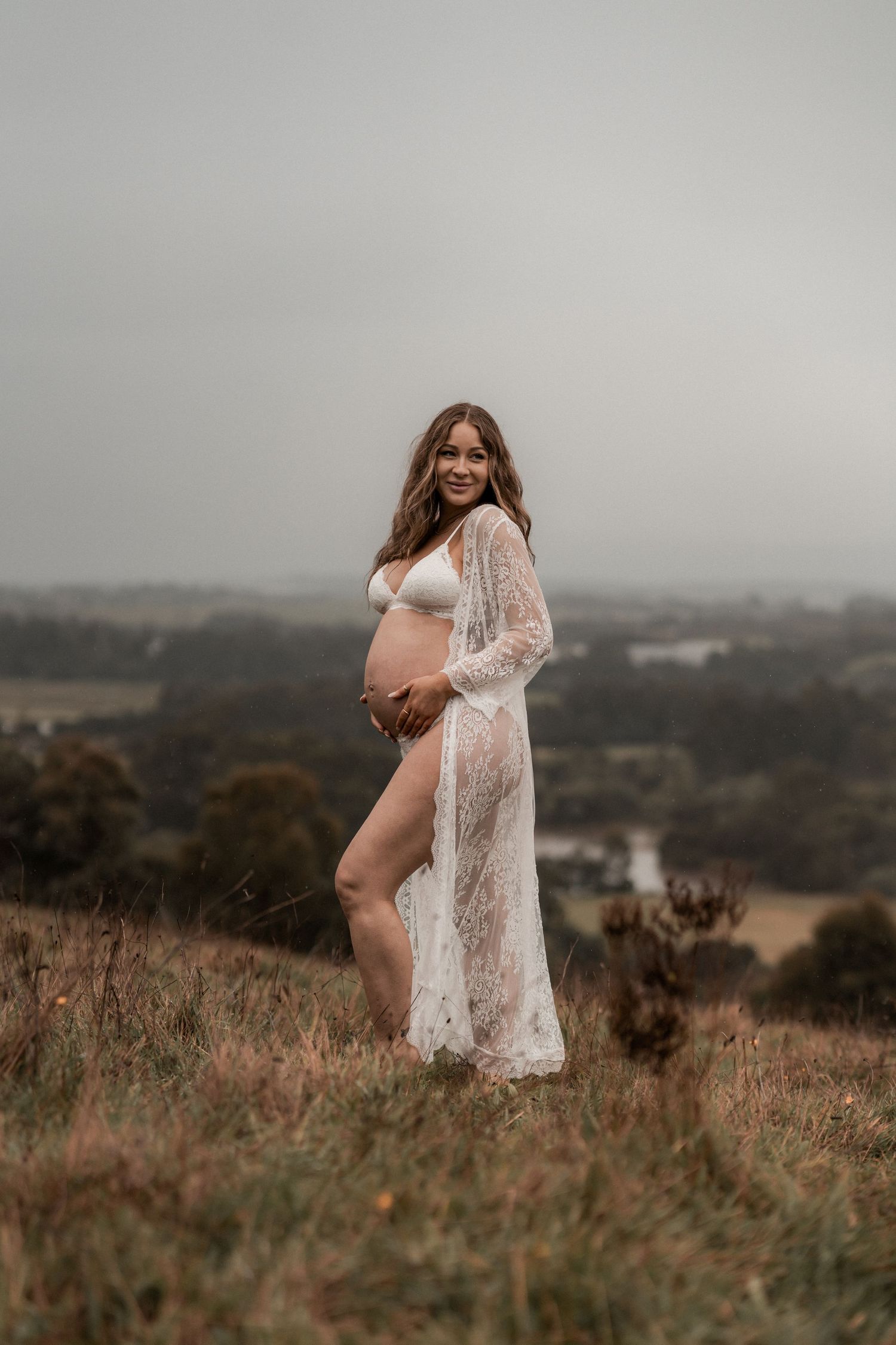 A pregnant figure in white lace robe poses on a hilltop against a misty countryside landscape at dusk.