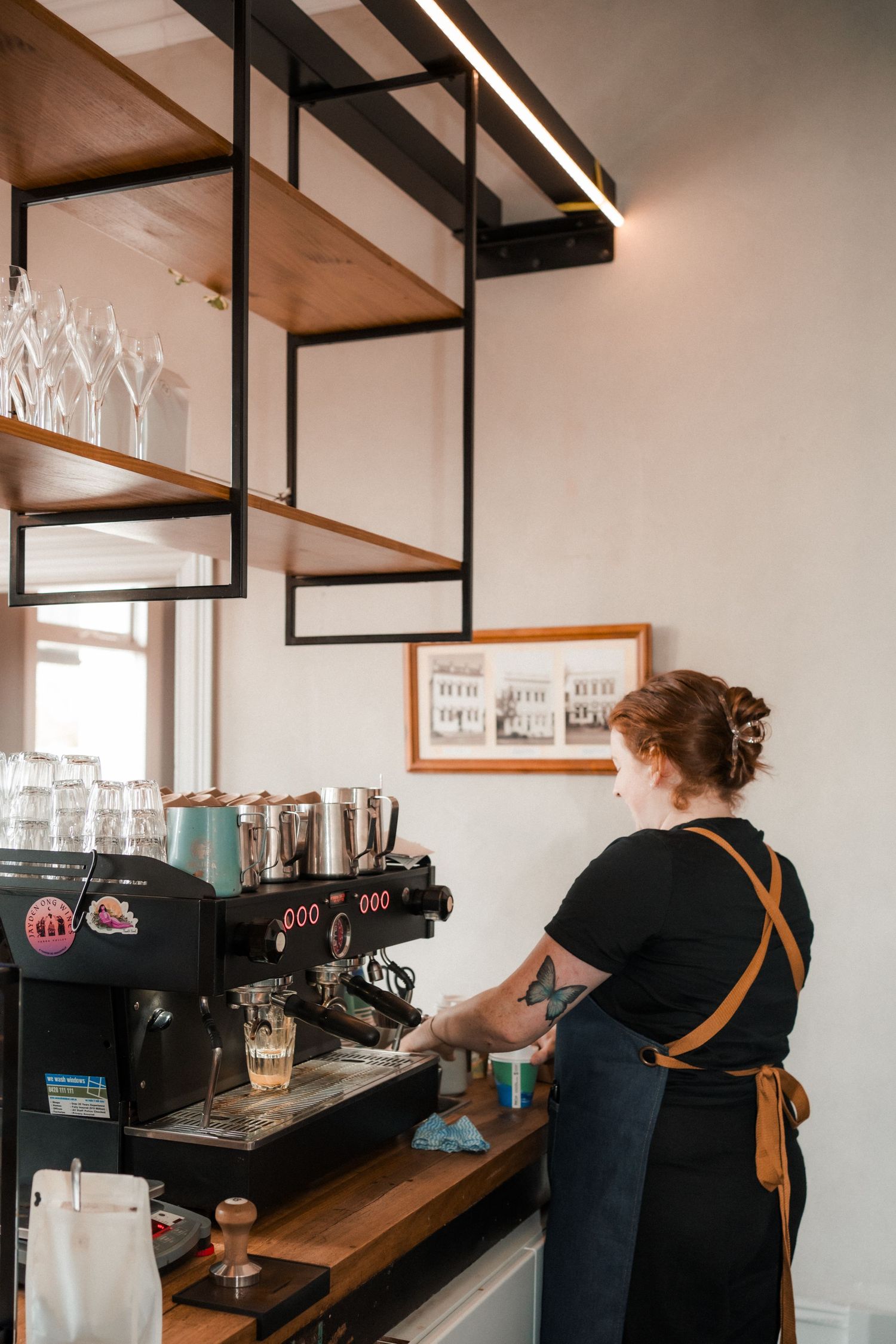 A barista in a black shirt working behind a wooden counter with industrial-style shelving and coffee equipment.