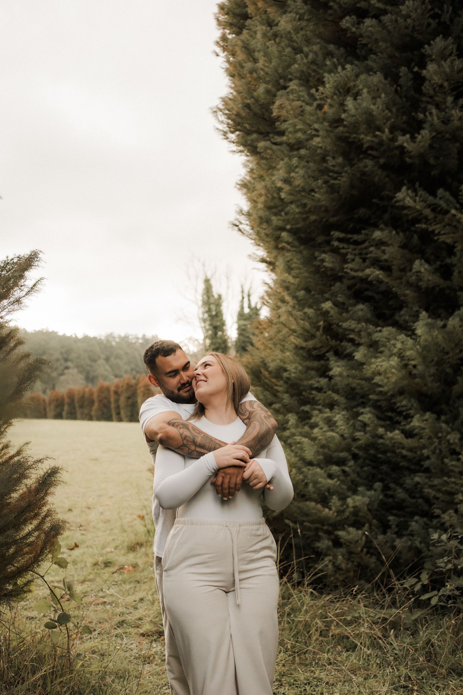 A couple shares a tender moment outdoors near evergreen trees during an autumn day photoshoot.