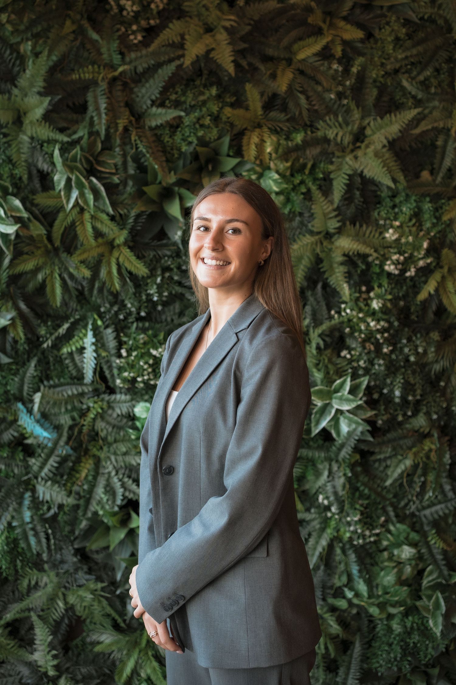 A person in a gray blazer poses against a lush green foliage wall background in multiple angles.