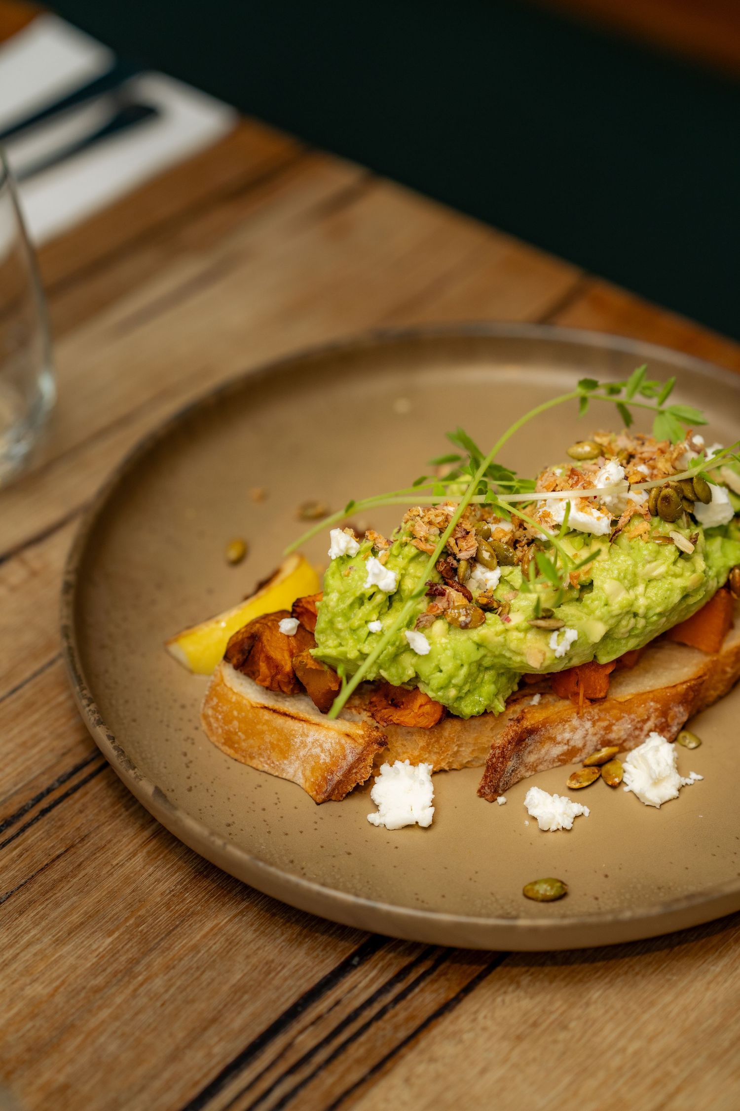 Grilled bread topped with avocado, crumbled feta cheese, and microgreens served on an elegant ceramic plate.