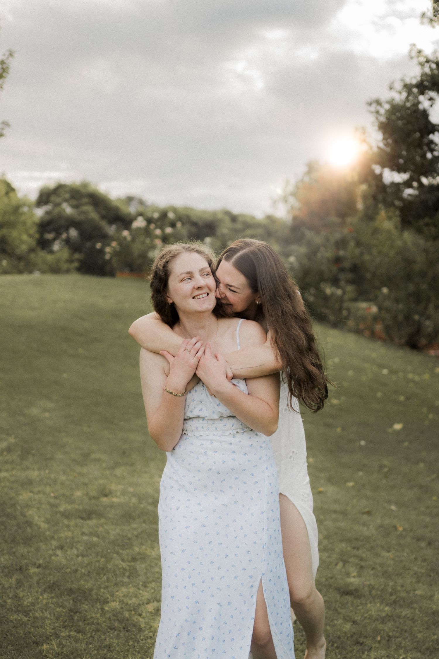 Two people in white dresses embrace and laugh joyfully together in a sunny outdoor field at sunset.