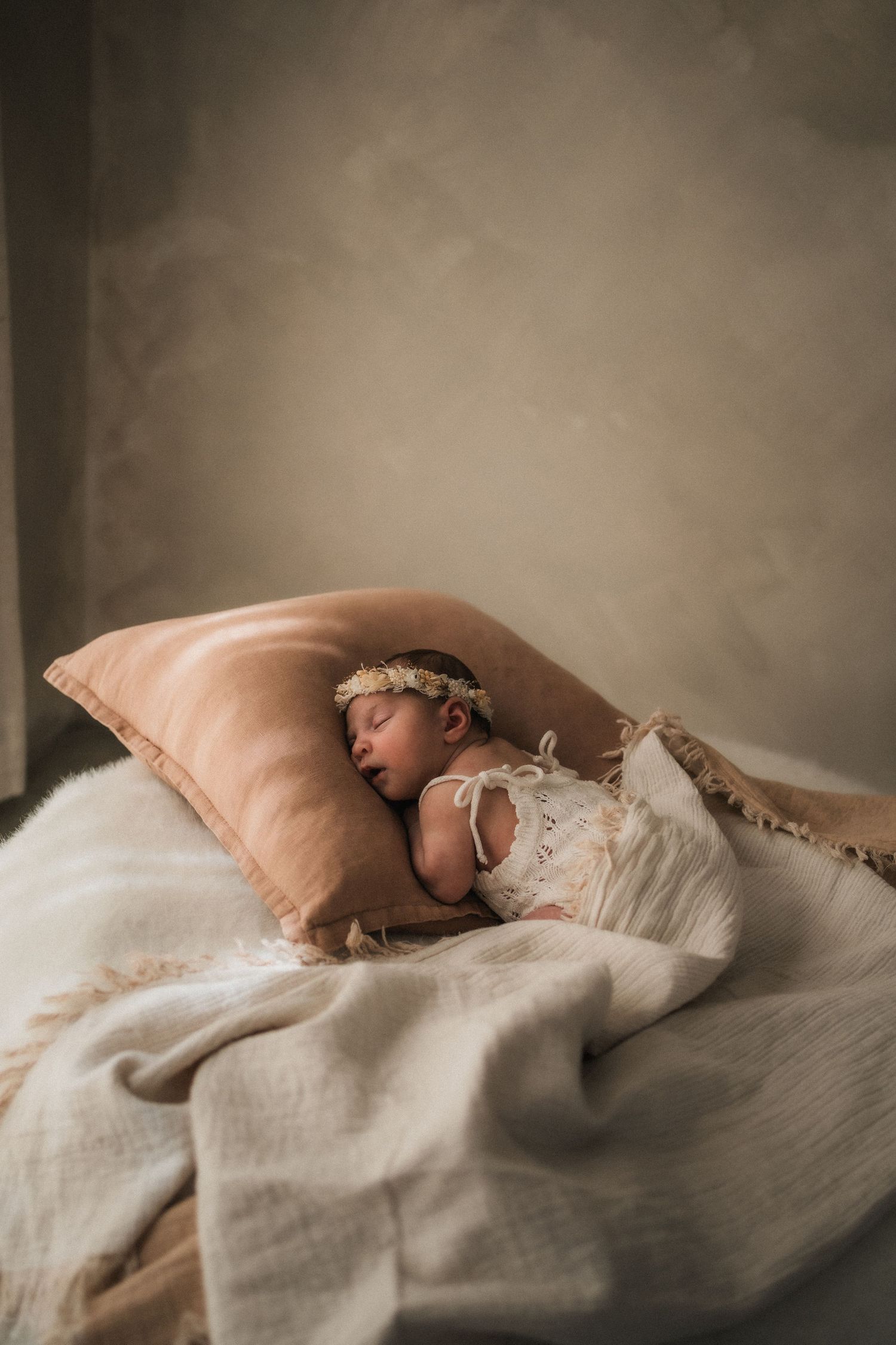 Newborn baby in a white lace romper and floral headband sleeping peacefully on a blush pillow with soft cream linen blankets.