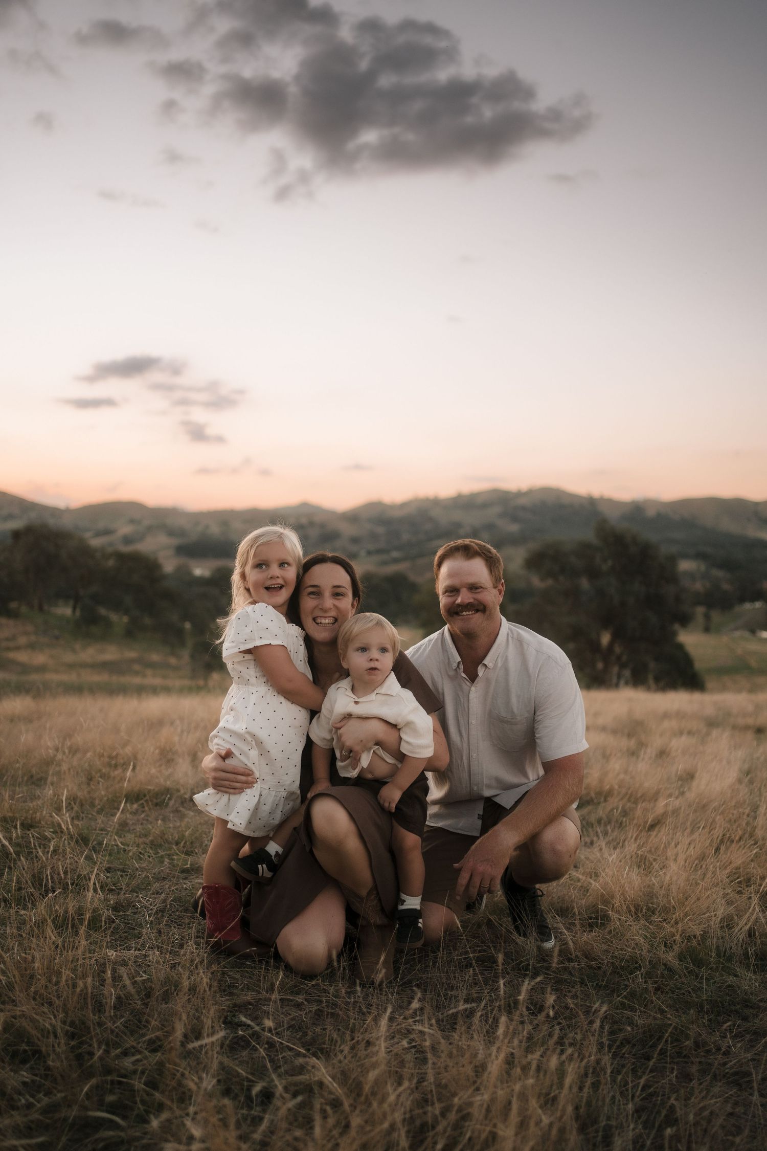 A family sits together in a golden field at sunset with mountains visible in the background.