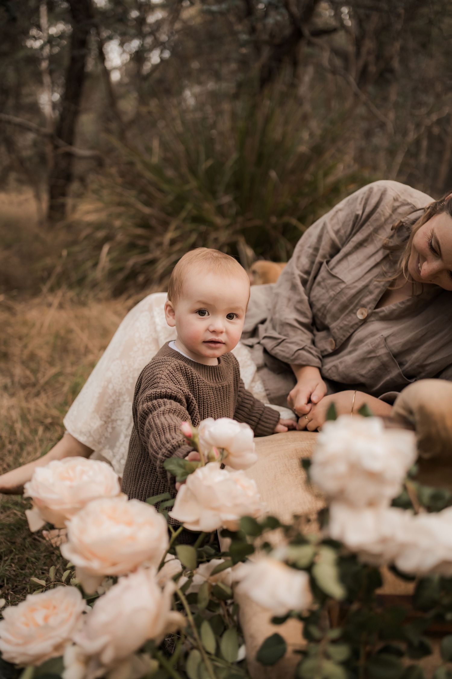 A small child in a brown sweater sits among blush-colored roses in a dreamy outdoor setting.