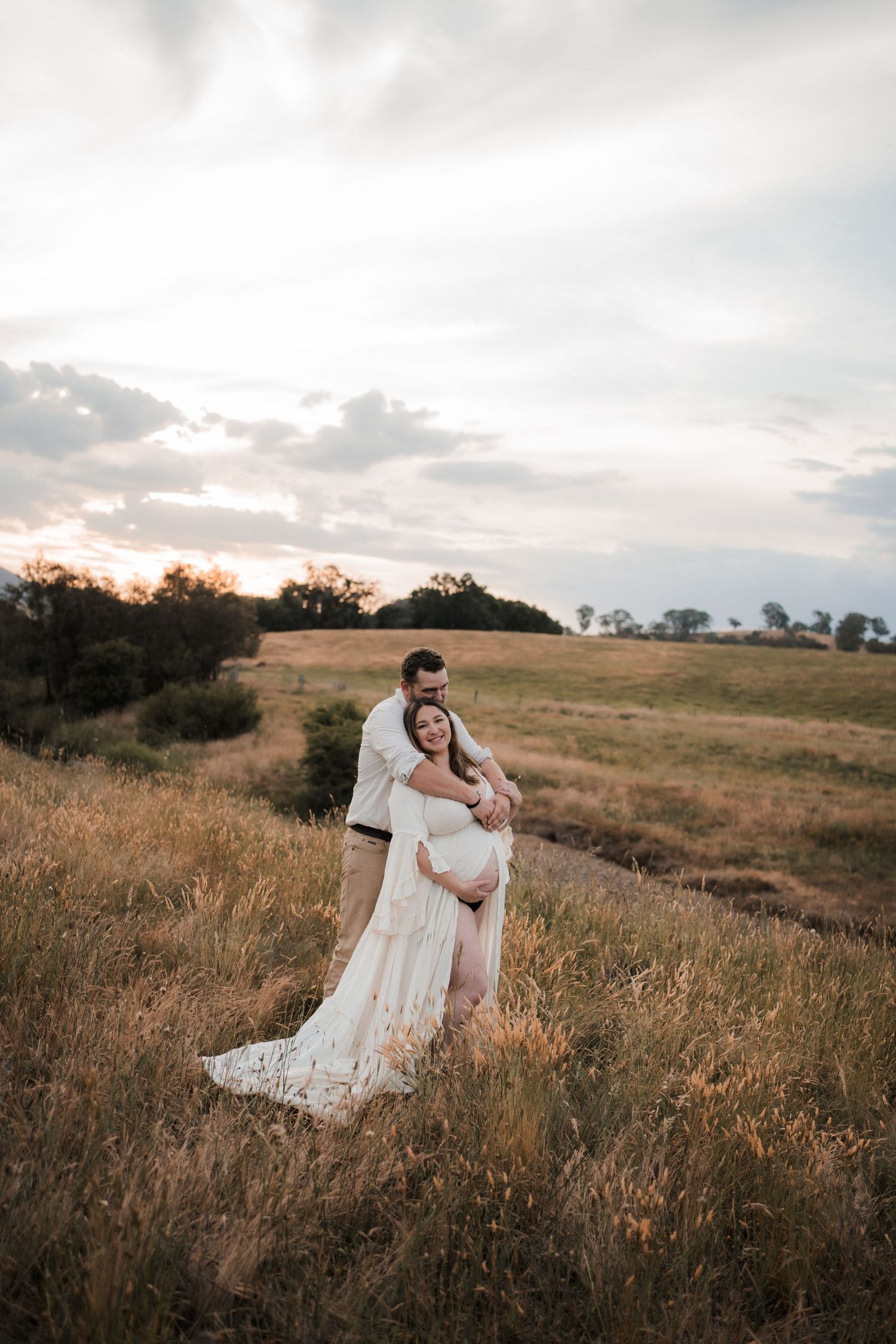 Couple embraces in romantic sunset scene on grassy hillside with dreamy golden hour lighting and soft clouds overhead.