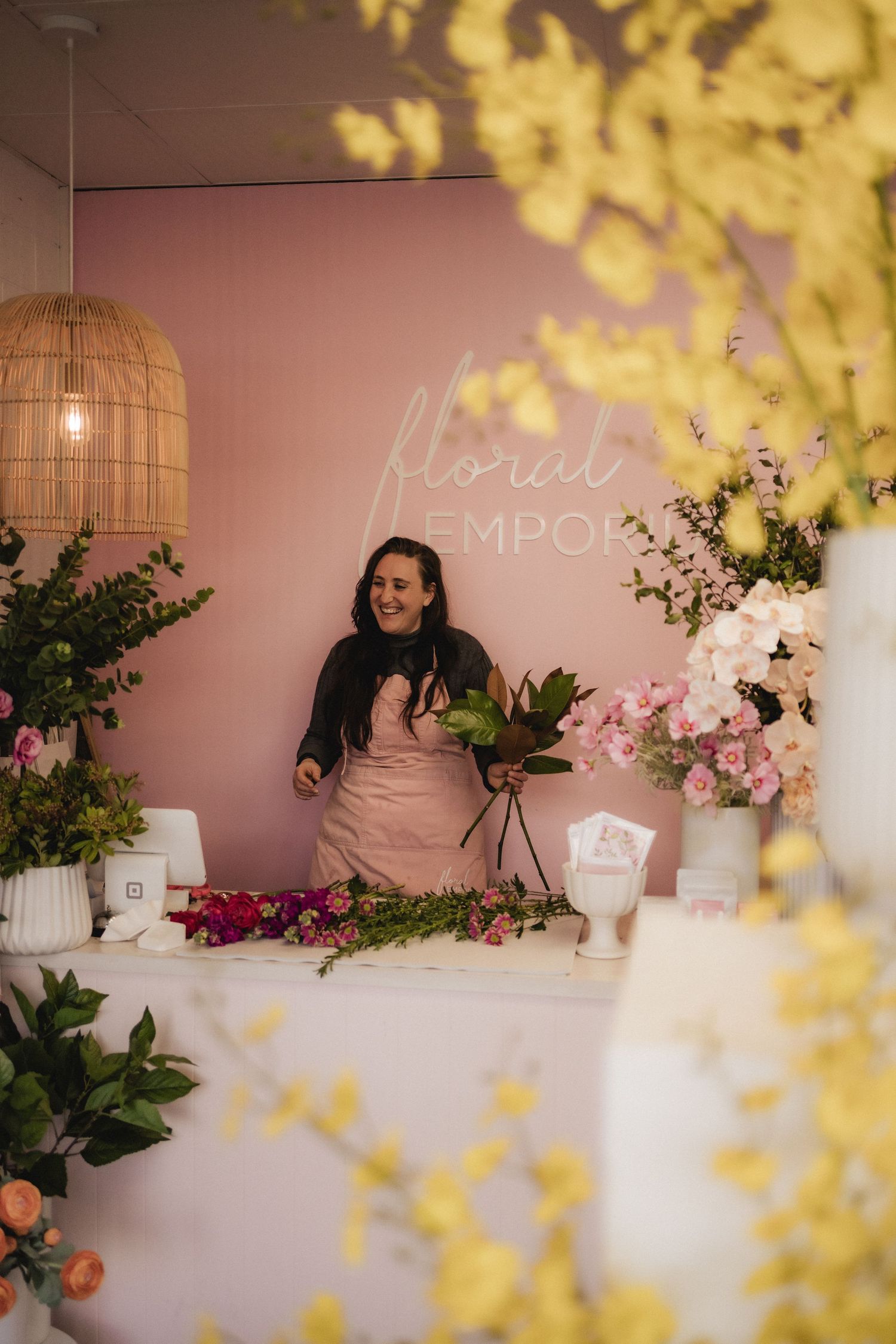 Floral workshop setup with pink walls, woven pendant light, and colorful flower arrangements on a white counter.