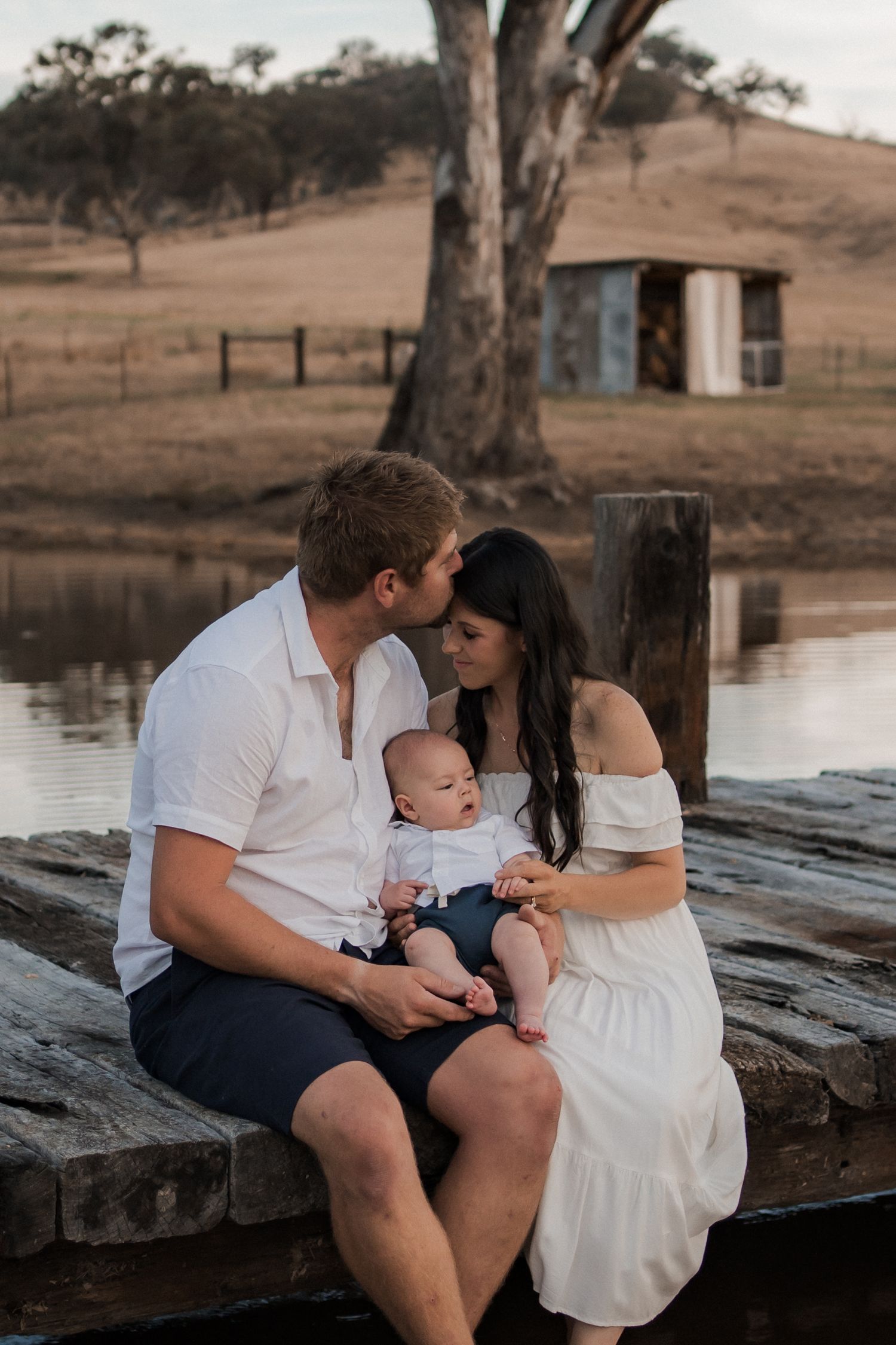 Family sitting together on wooden dock by a pond with rustic shed and gum trees in background.